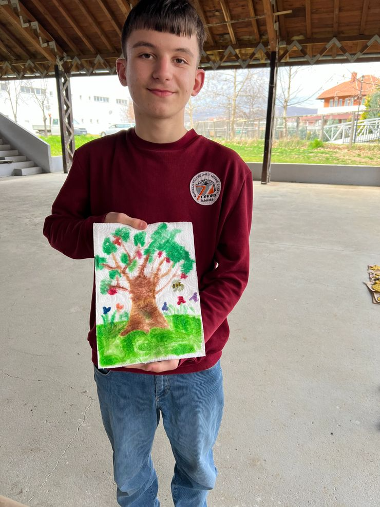 Smiling boy in a maroon long-sleeve shirt holding a colorful drawing of a large tree with green leaves, a brown trunk, and various small colorful flowers on a grassy hill, under a wooden shelter with outdoor area in the background.