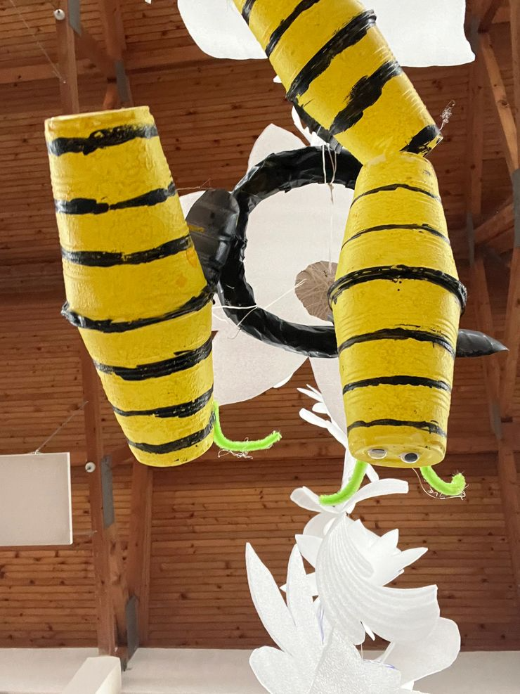 A hanging decoration featuring a black and yellow striped bee with green pipe cleaner antennae and googly eyes, attached to white paper flower decorations.