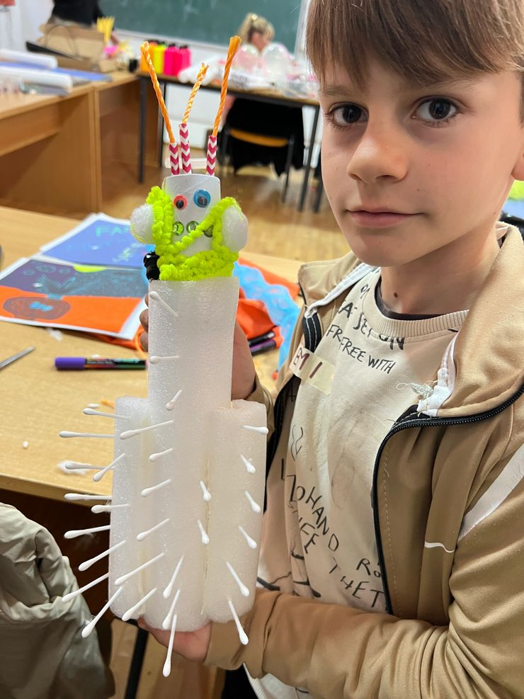 A young boy is holding a craft project resembling a small robot or figure made from foam, pipe cleaners, googly eyes, and candy sticks, in a classroom setting.