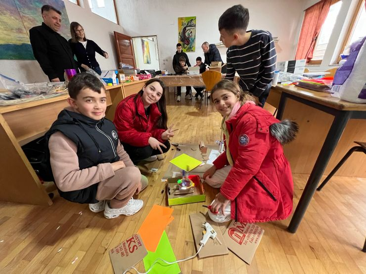 Children working on a science project in a classroom, with adults in the background, colorful papers, and electronic components.