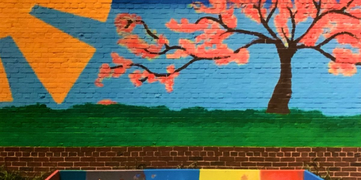 Colorful mural of a tree with pink blossoms on a brick wall, with blue, yellow, and red geometric shapes on the left.
