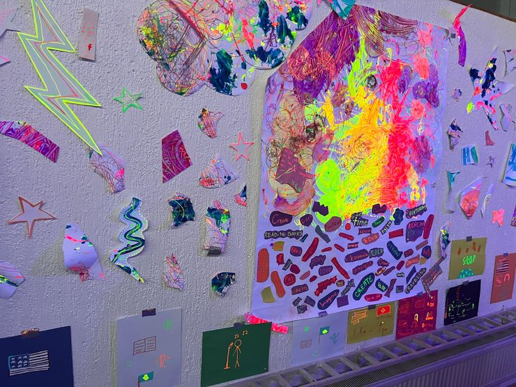 Colorful, abstract artwork on a wall with torn paper shapes, stickers, and glow-in-the-dark elements.