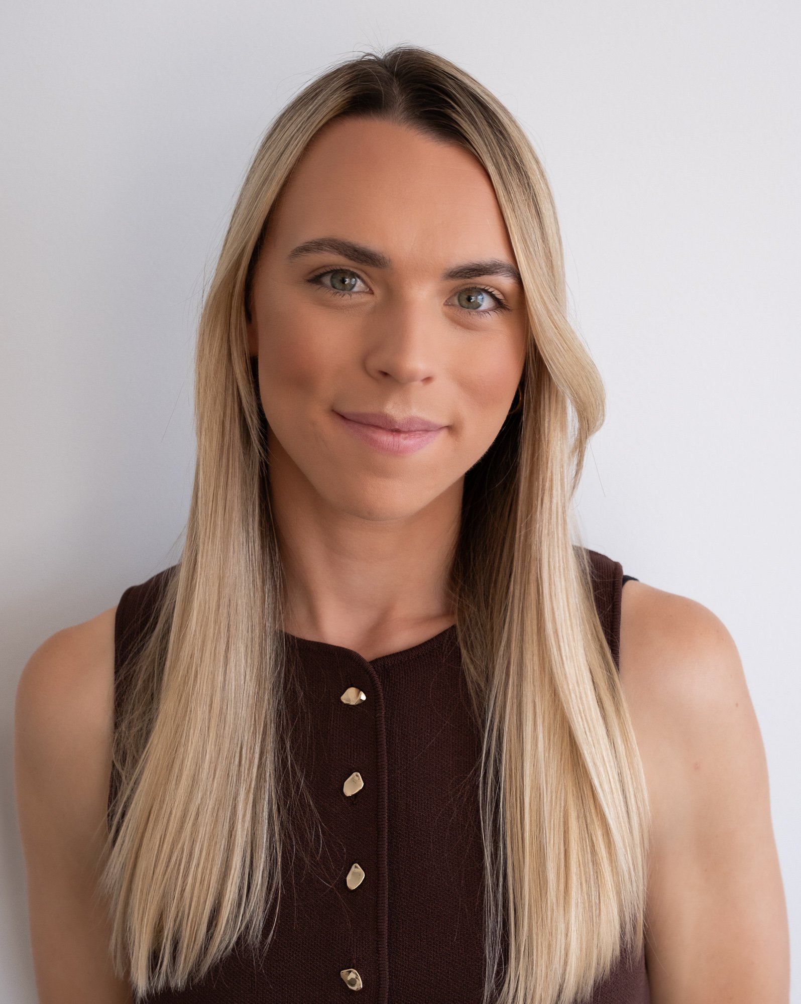 A woman with long blonde hair, light skin, and green eyes, wearing a sleeveless dark brown top with gold button details, smiling softly against a plain white background.