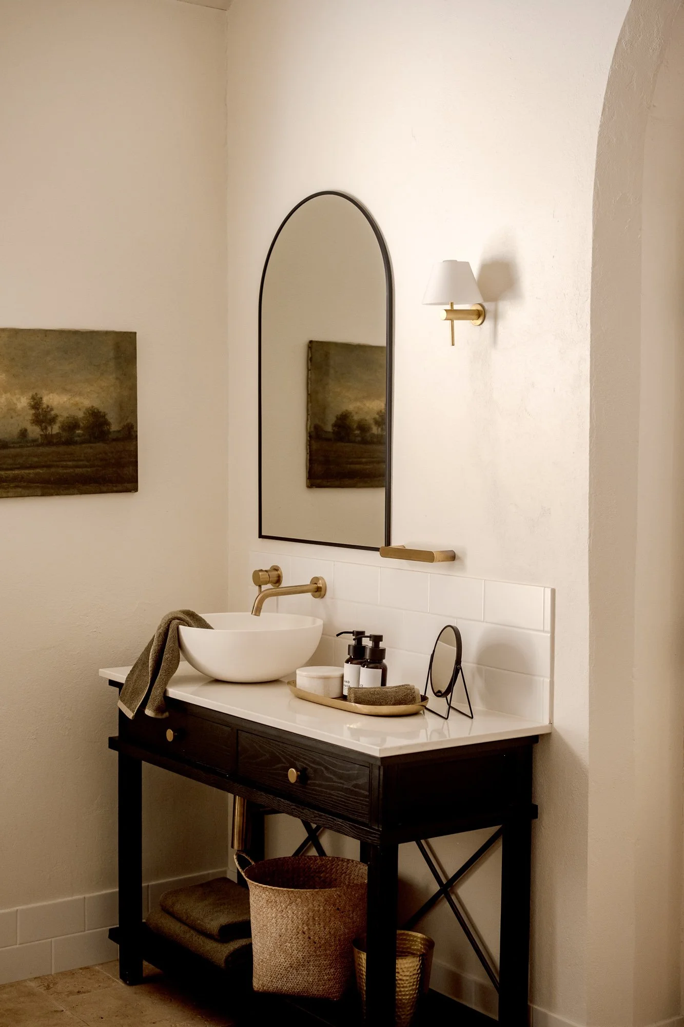A modern bathroom vanity with a white vessel sink, black wooden table, and a large arched mirror. There are soap dispensers, a small mirror, and towels on the countertop. A wall sconce light is mounted to the right of the arched mirror.