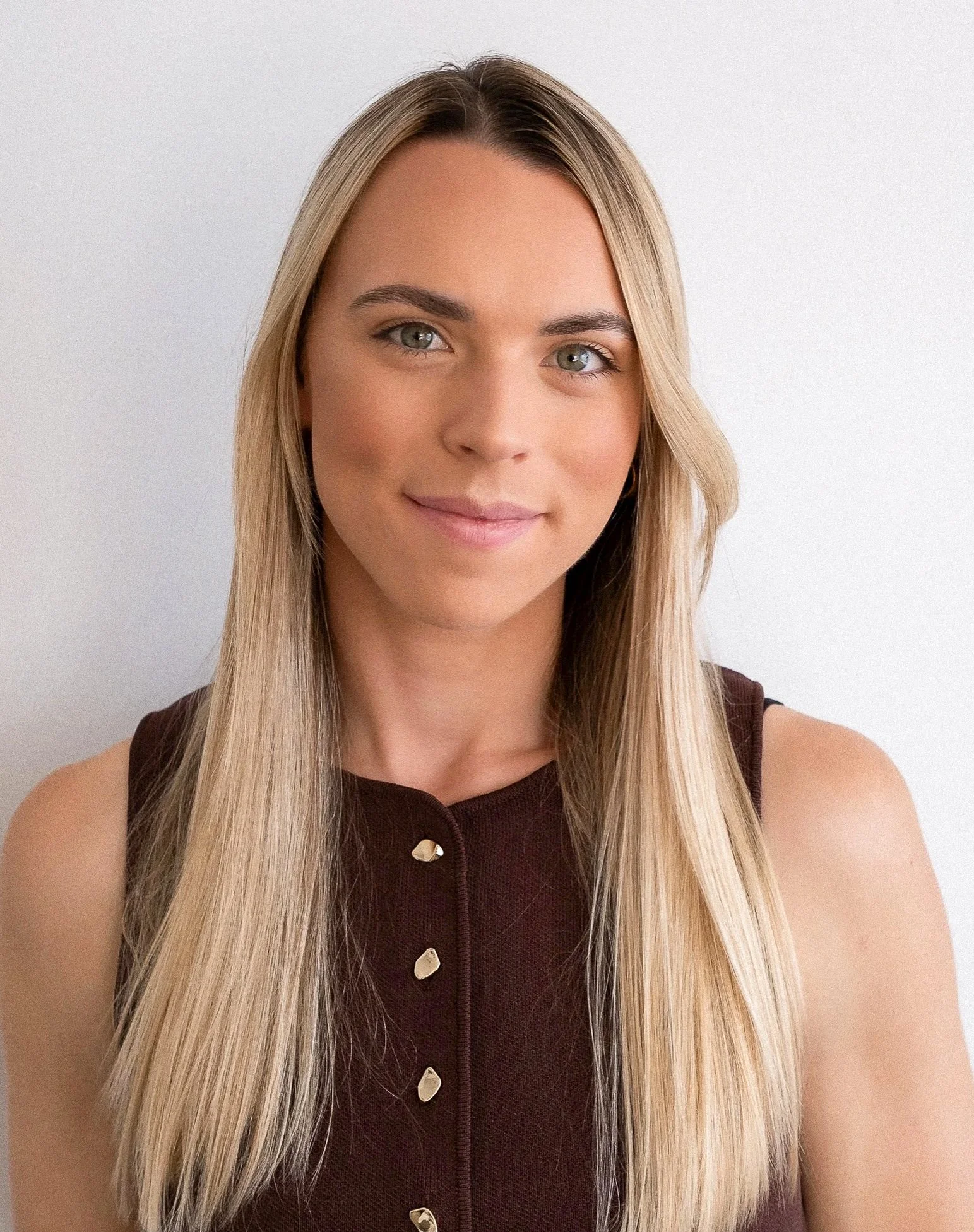 A young woman with long blonde hair, wearing a sleeveless dark brown top with gold buttons, smiling softly against a plain light gray background.