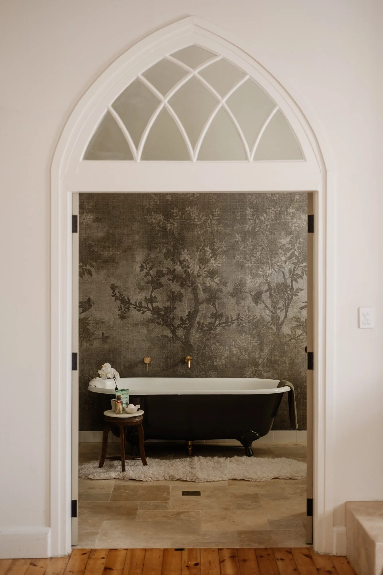 Bathroom with a black clawfoot tub, decorative wallpaper, with a tree pattern, and a small wooden stool with items on it, viewed through an arched doorway.