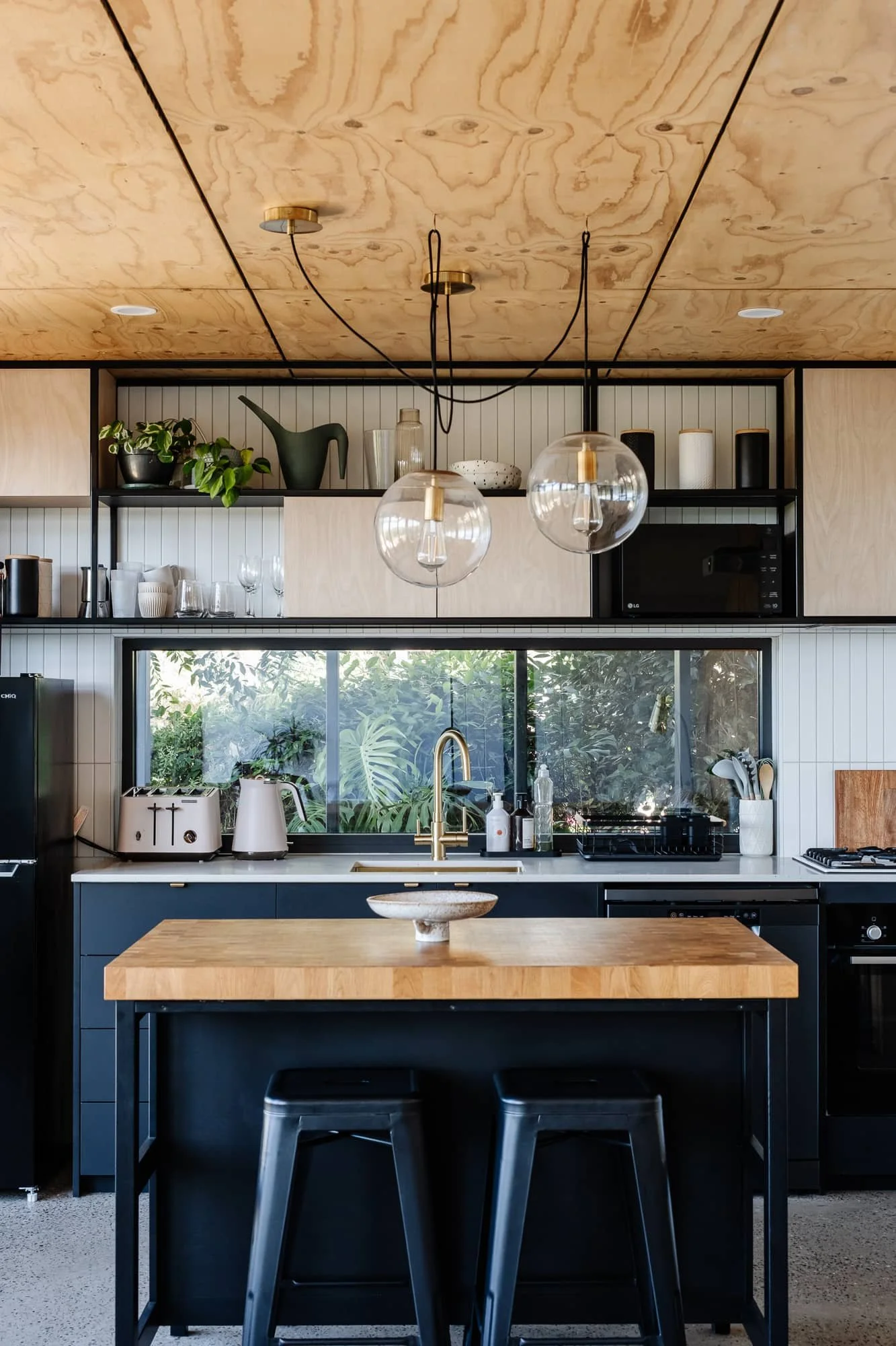 Modern kitchen with wooden ceiling, black-finished cabinets, gold faucet, and a large window with greenery outside. Features include a kitchen island, pendant lights, shelves with decor, and various kitchen appliances.