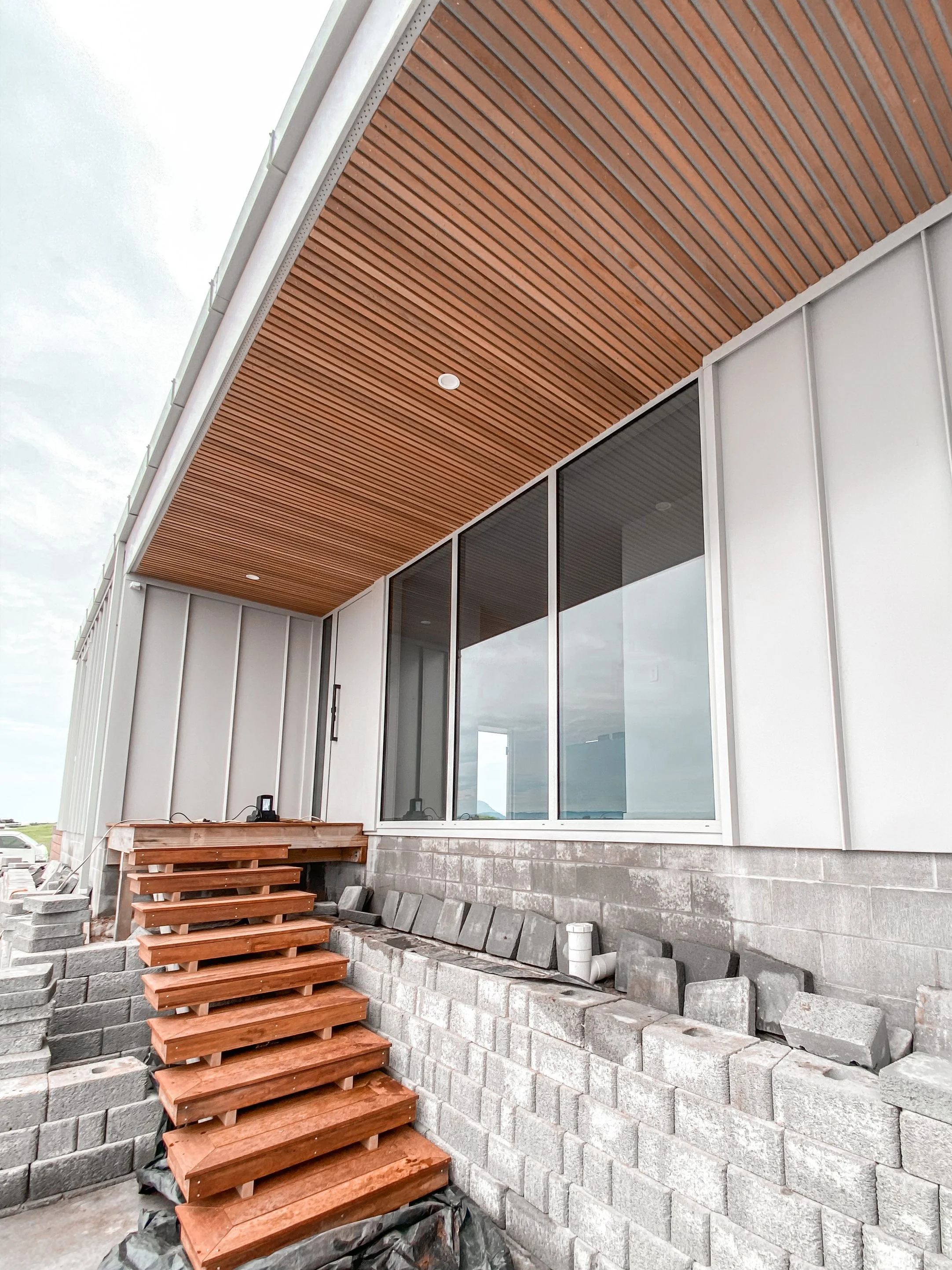 Exterior of a modern building under construction, showing a set of wooden stairs leading to a small deck, large windows, and a wooden ceiling overhang.