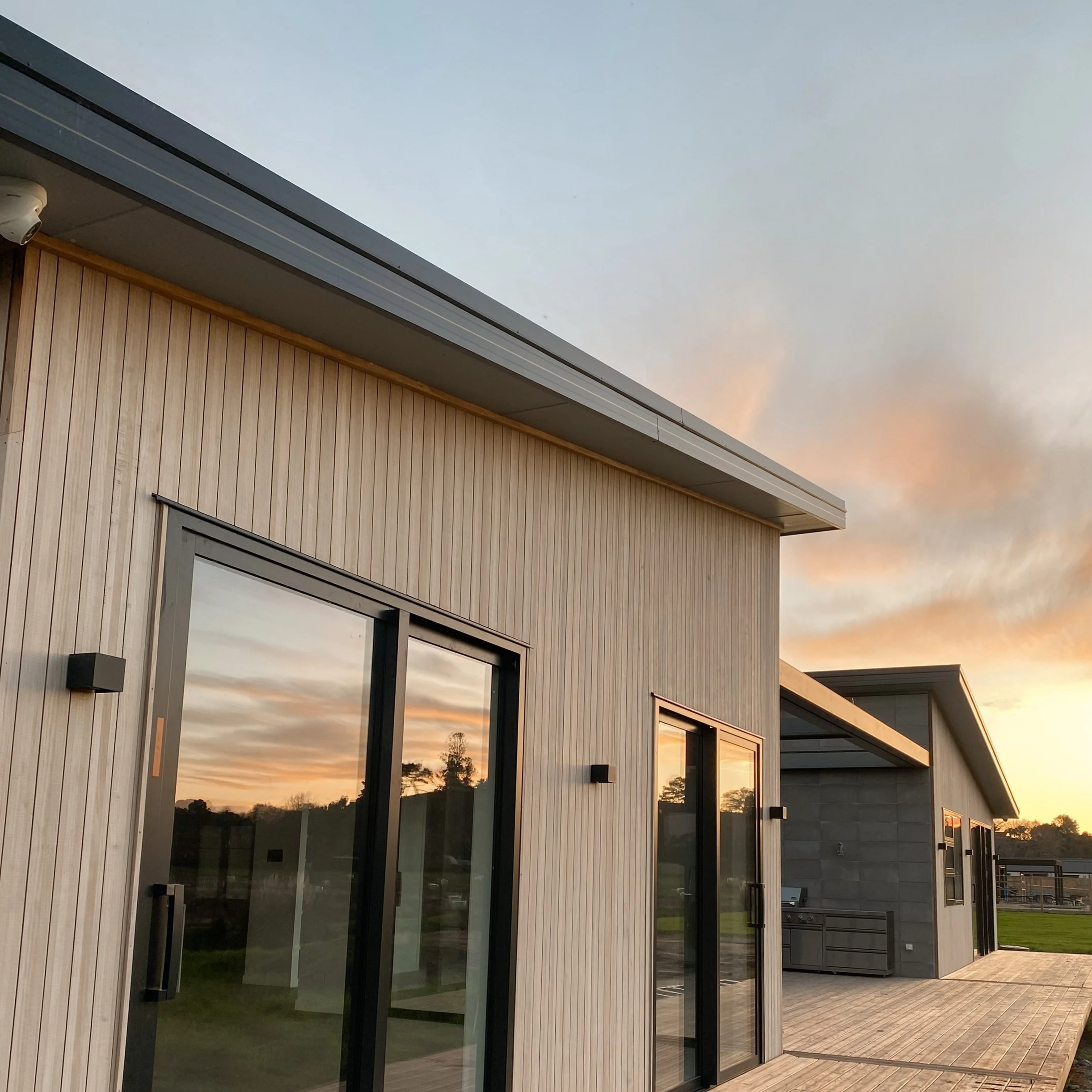 Modern building with vertical wood siding and large glass sliding doors at sunset, with a wooden deck and outdoor space.