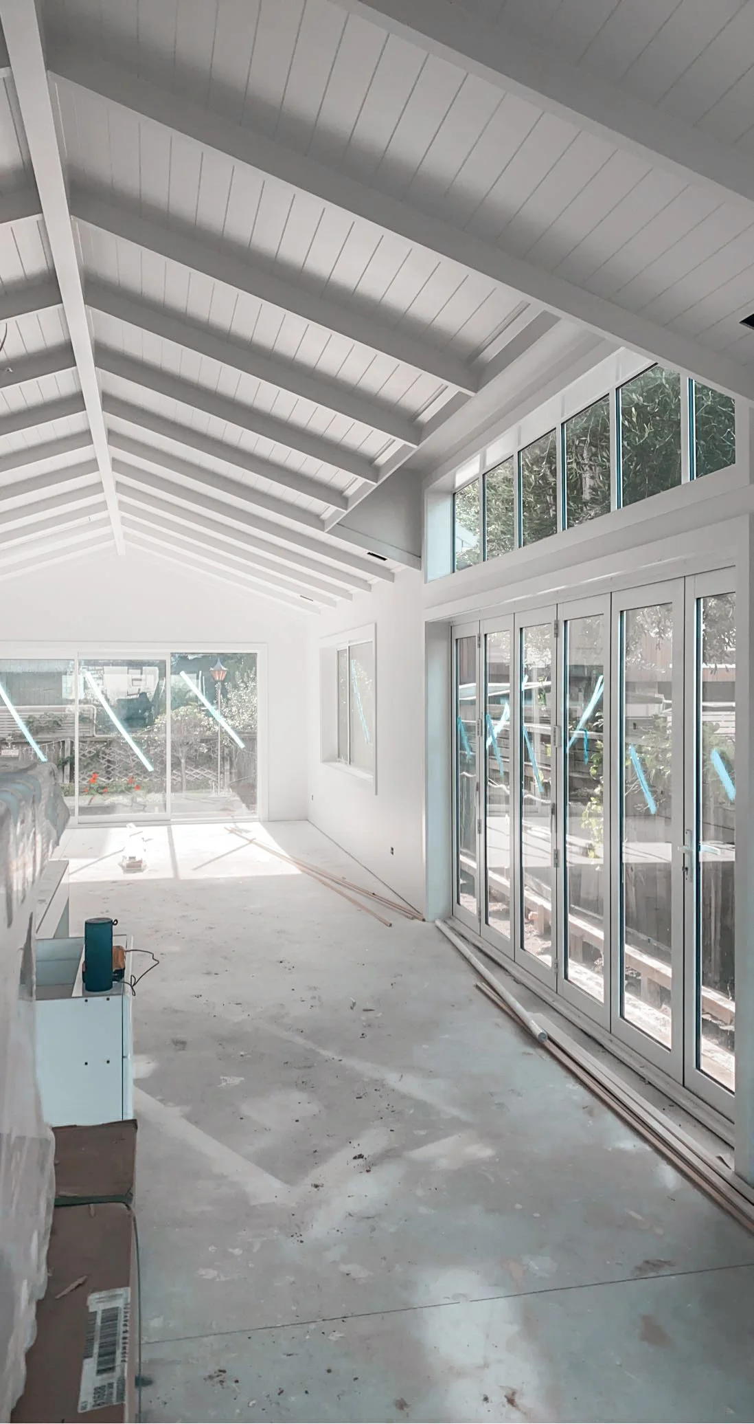 Interior of a house under construction with large sliding glass doors, high vaulted ceiling with exposed white beams, and a partially finished concrete floor.