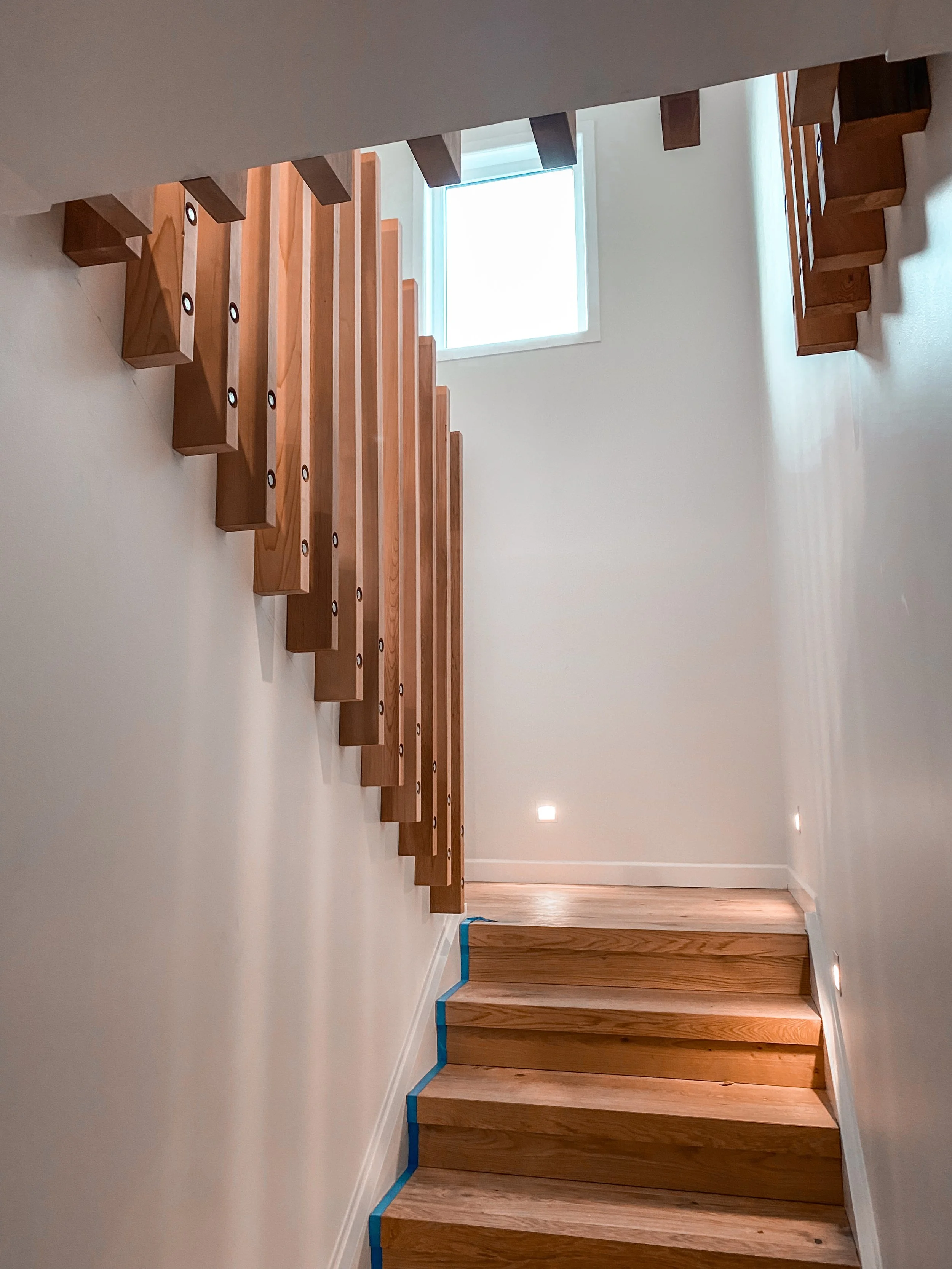 Interior view of a wooden staircase in a well-lit space with a window at the top, showing steps and handrails.
