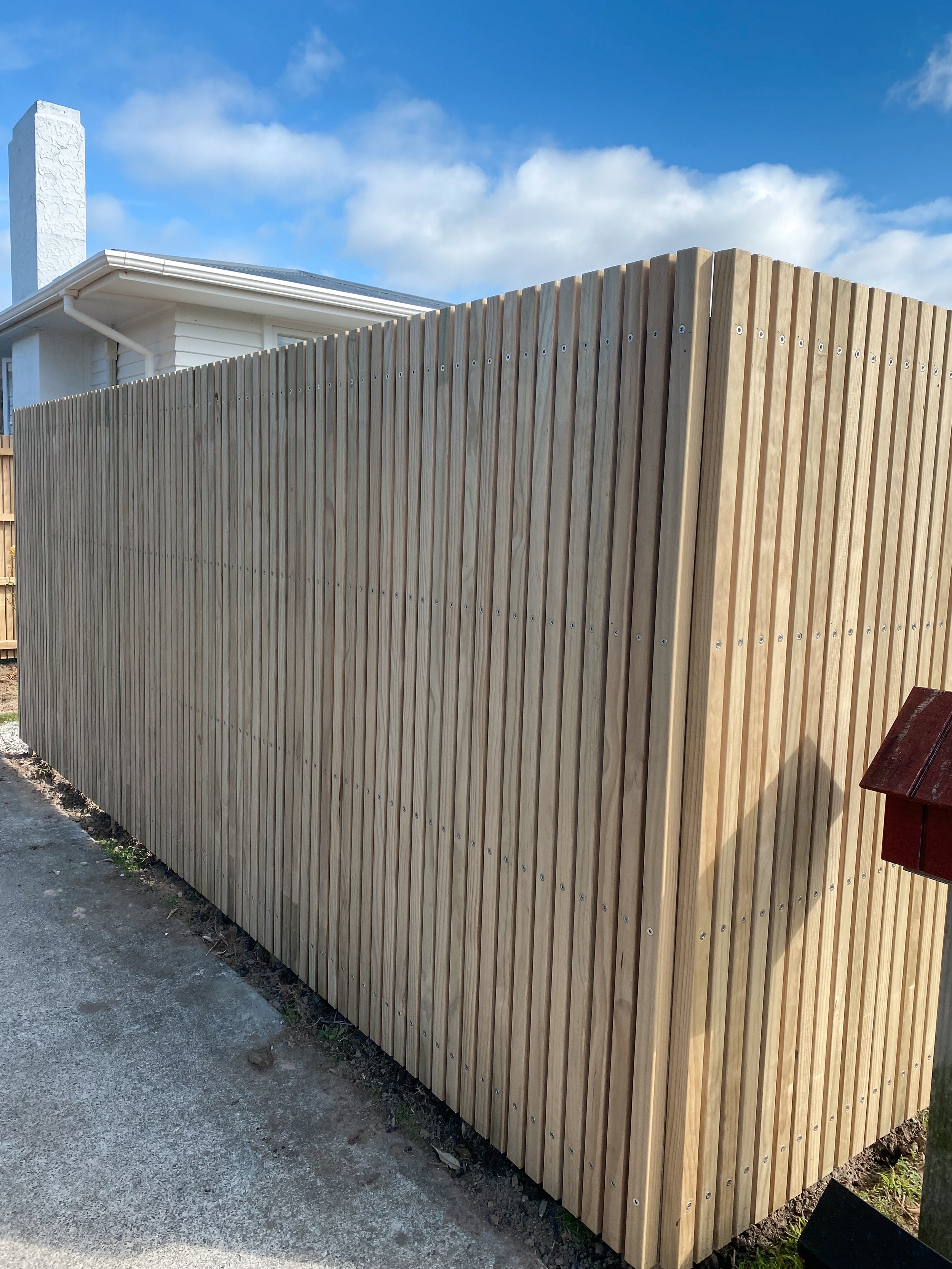 A newly constructed wooden privacy fence with vertical slats and metal screws, next to a concrete sidewalk, with a white house and blue sky in the background.