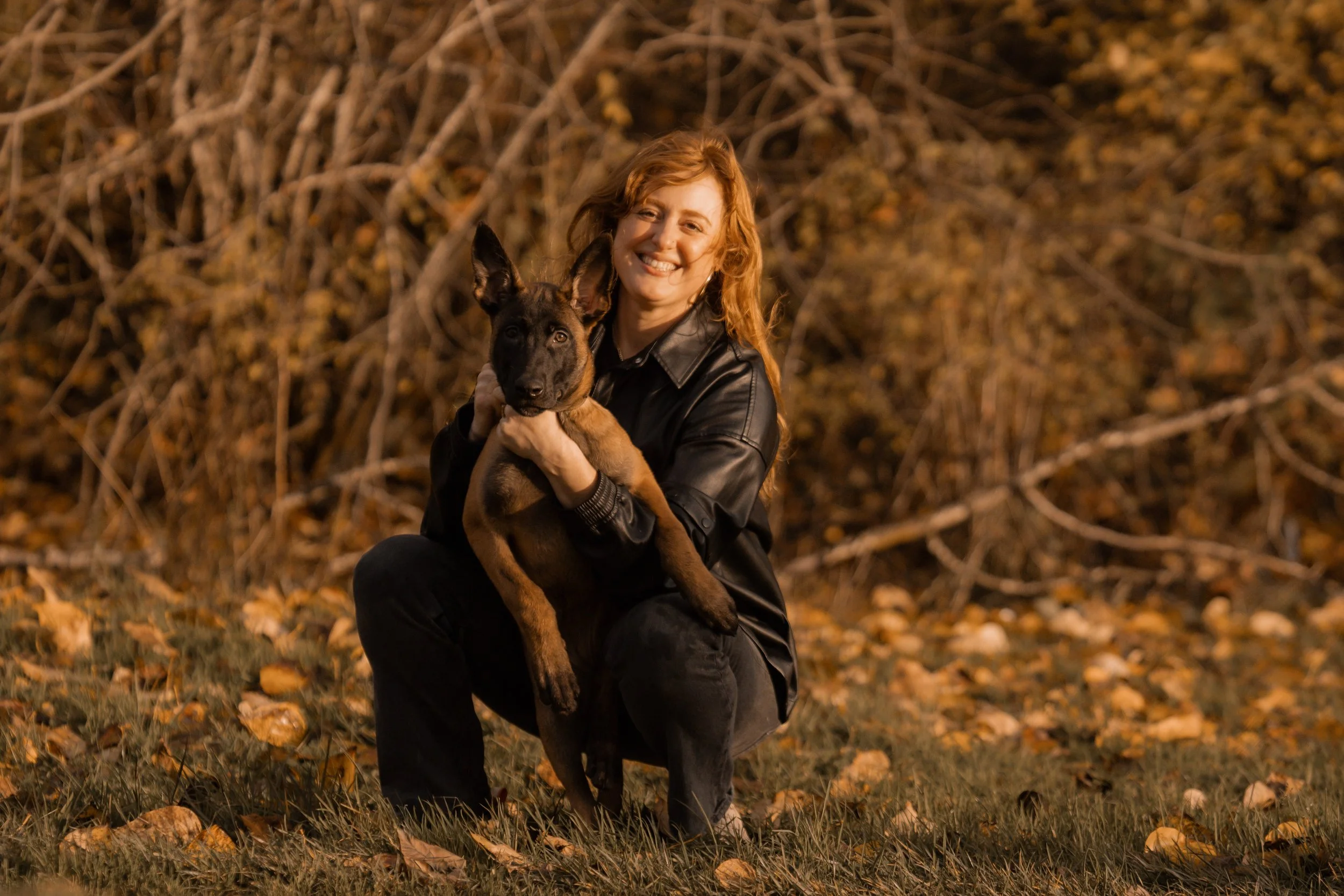 A woman with red hair wearing a black leather jacket kneeling outdoors with a young black and tan German Shepherd puppy in her arms, surrounded by fallen autumn leaves and trees in the background.