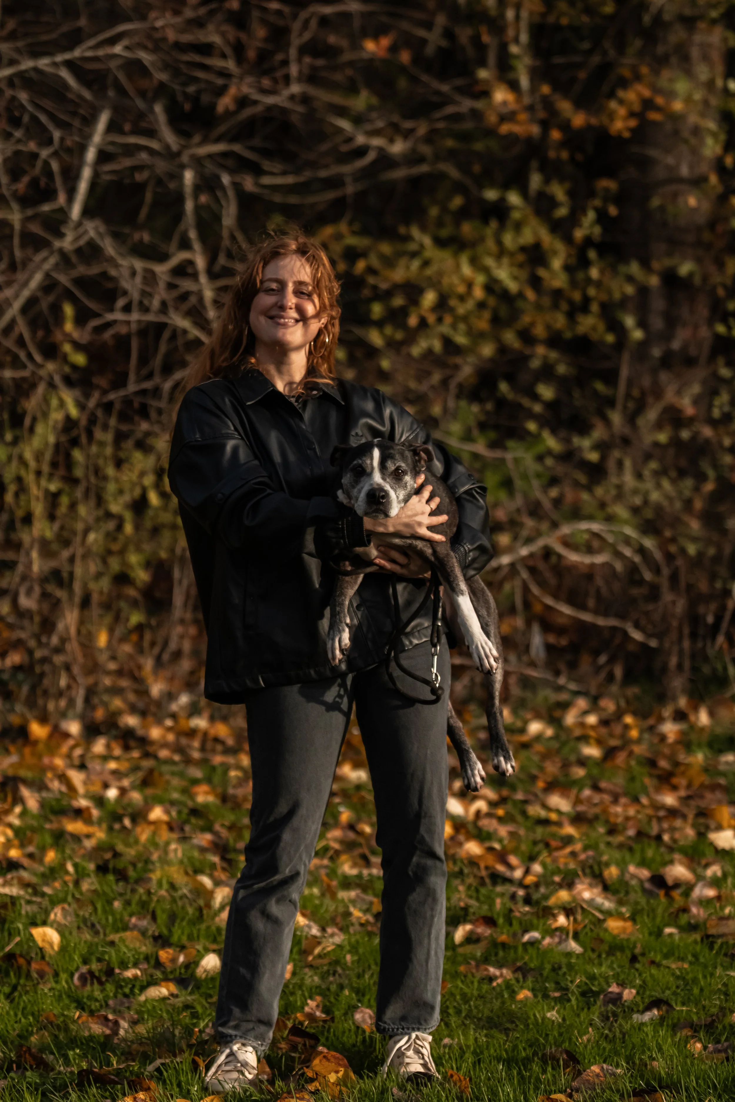 A woman with curly red hair, wearing a black jacket, smiling and holding a black and white dog in an outdoor setting with autumn leaves on the ground and trees in the background.
