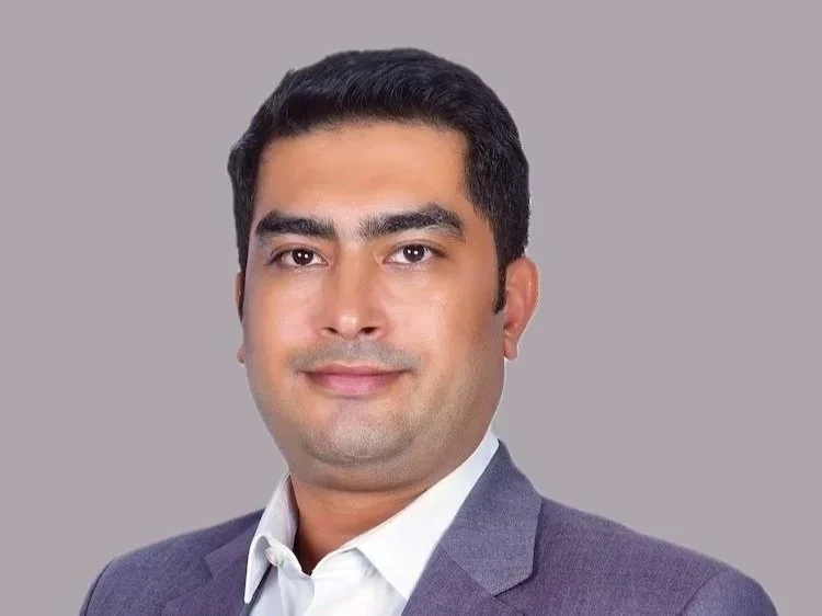 Smiling man with dark hair, glasses, and a black shirt against a plain light background.