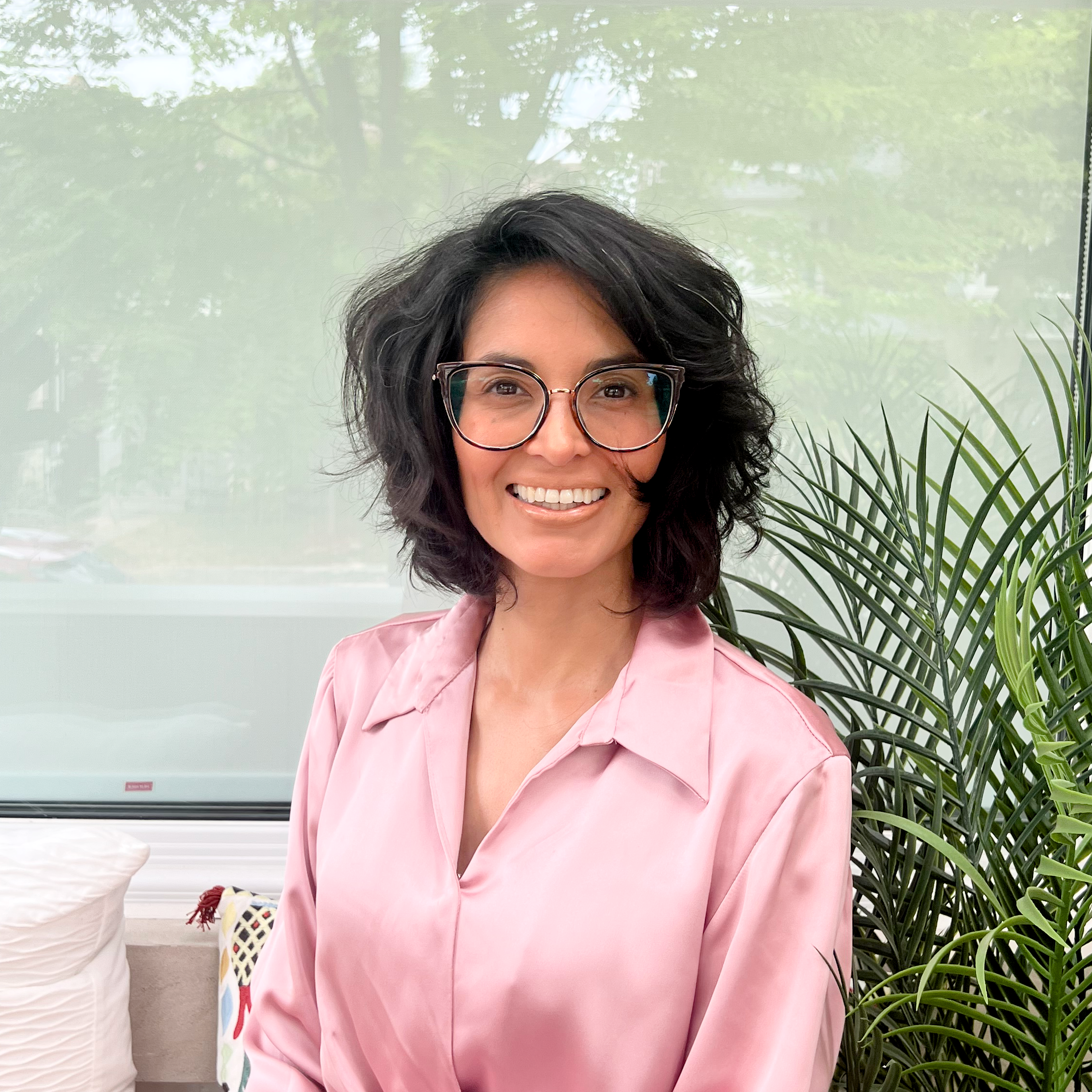 A woman with short, curly black hair wearing glasses and a pink satin blouse, smiling, sitting indoors near a large green plant with a window and trees visible outside.