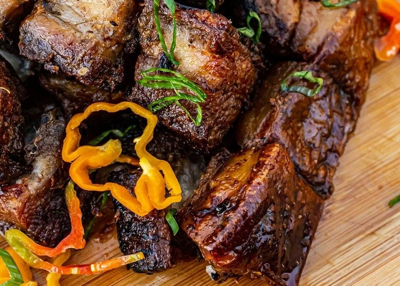 Close-up of cooked beef short ribs garnished with green herbs and sliced peppers on a wooden cutting board.