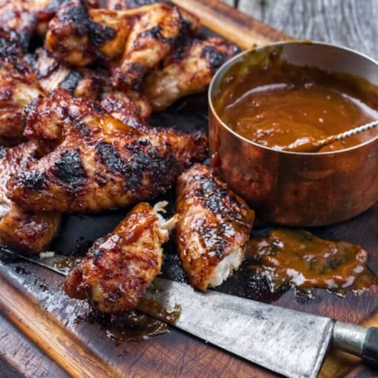 Grilled chicken wings with barbecue sauce on a wooden cutting board, alongside a small copper bowl of barbecue sauce and a knife.