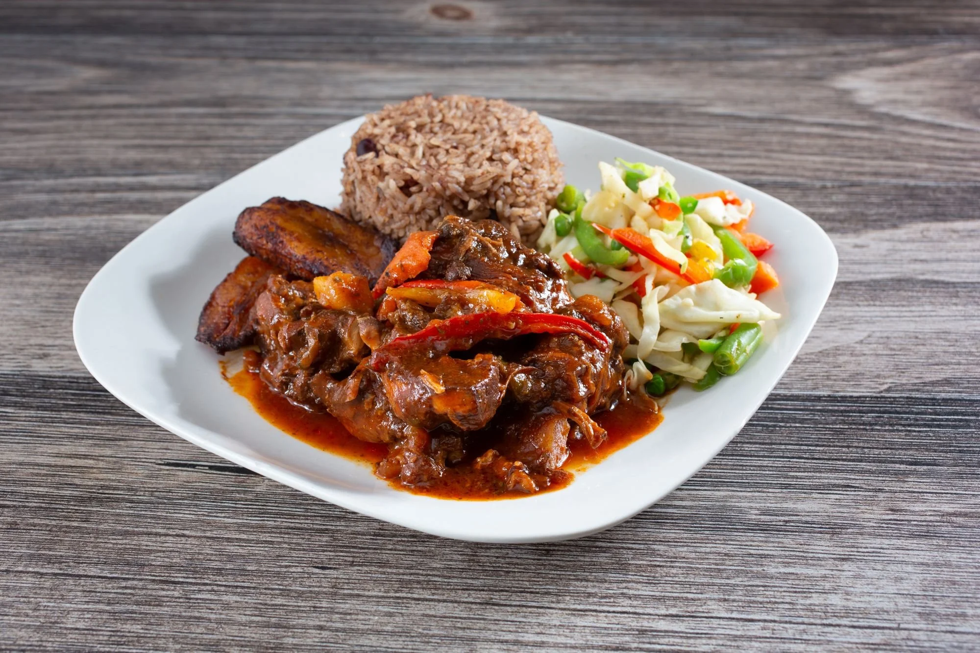 Plate of Jamaican cuisine with rice and peas, fried plantains, beef stew, and jamaican coleslaw on a wooden table.