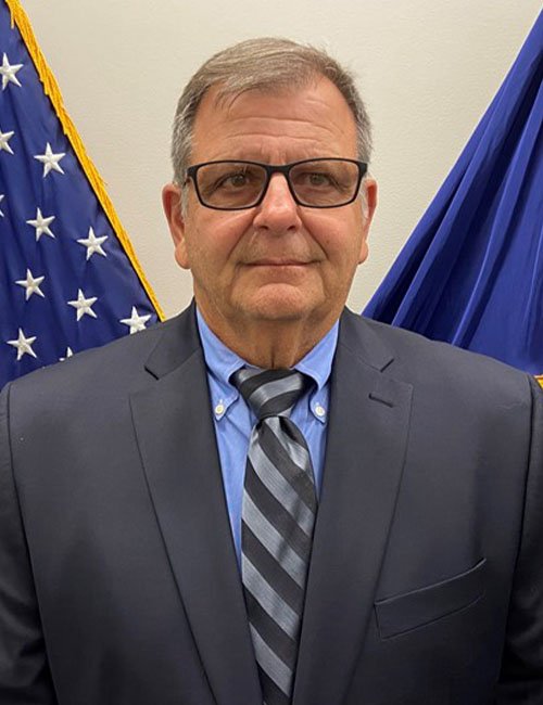 A man wearing glasses, a dark suit, and a striped tie standing in front of American flags.