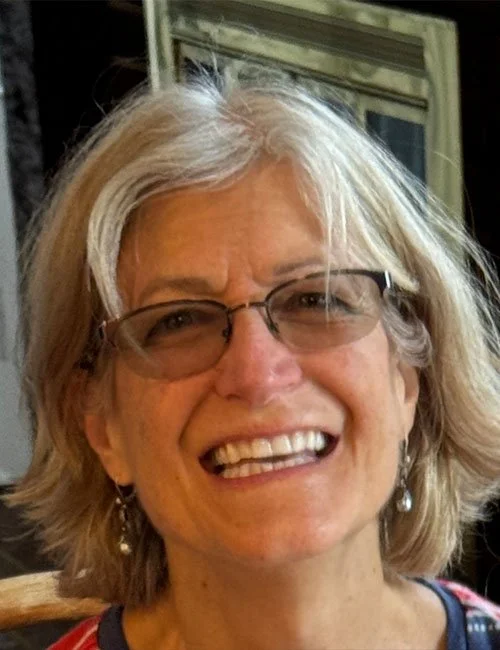 A smiling middle-aged woman with glasses and earrings, with light-colored hair, in an indoor setting.