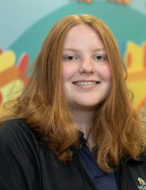 A young woman with long, wavy red hair smiling in front of a colorful, abstract background.