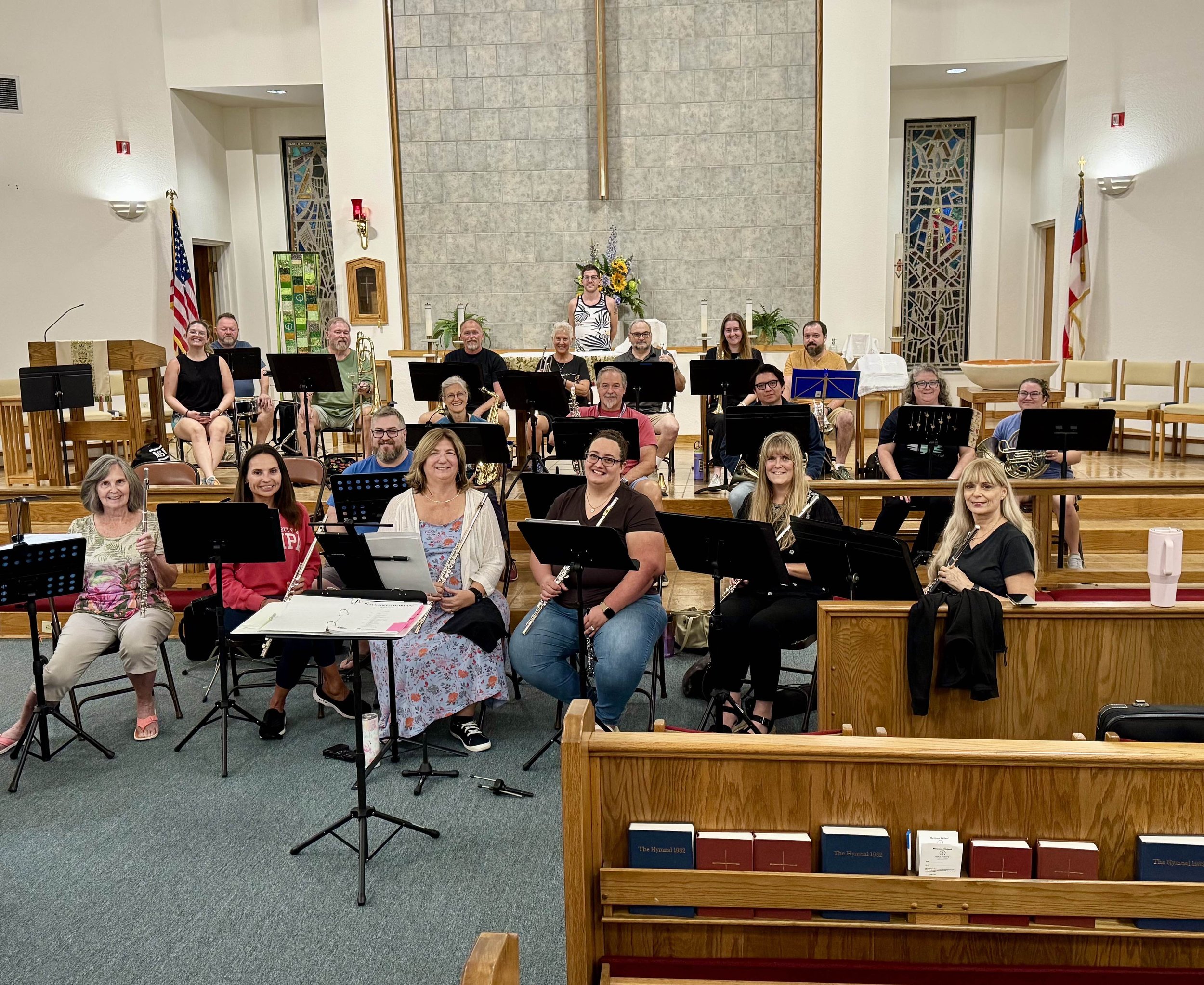 A group of musicians with various instruments posing inside a church or concert hall. They are sitting and standing on a wooden stage with an altar behind them, decorated with flowers and candles. Flags are on either side near the walls.