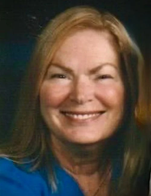 Close-up of a woman with shoulder-length brown hair, smiling, wearing a blue top, against a dark background.