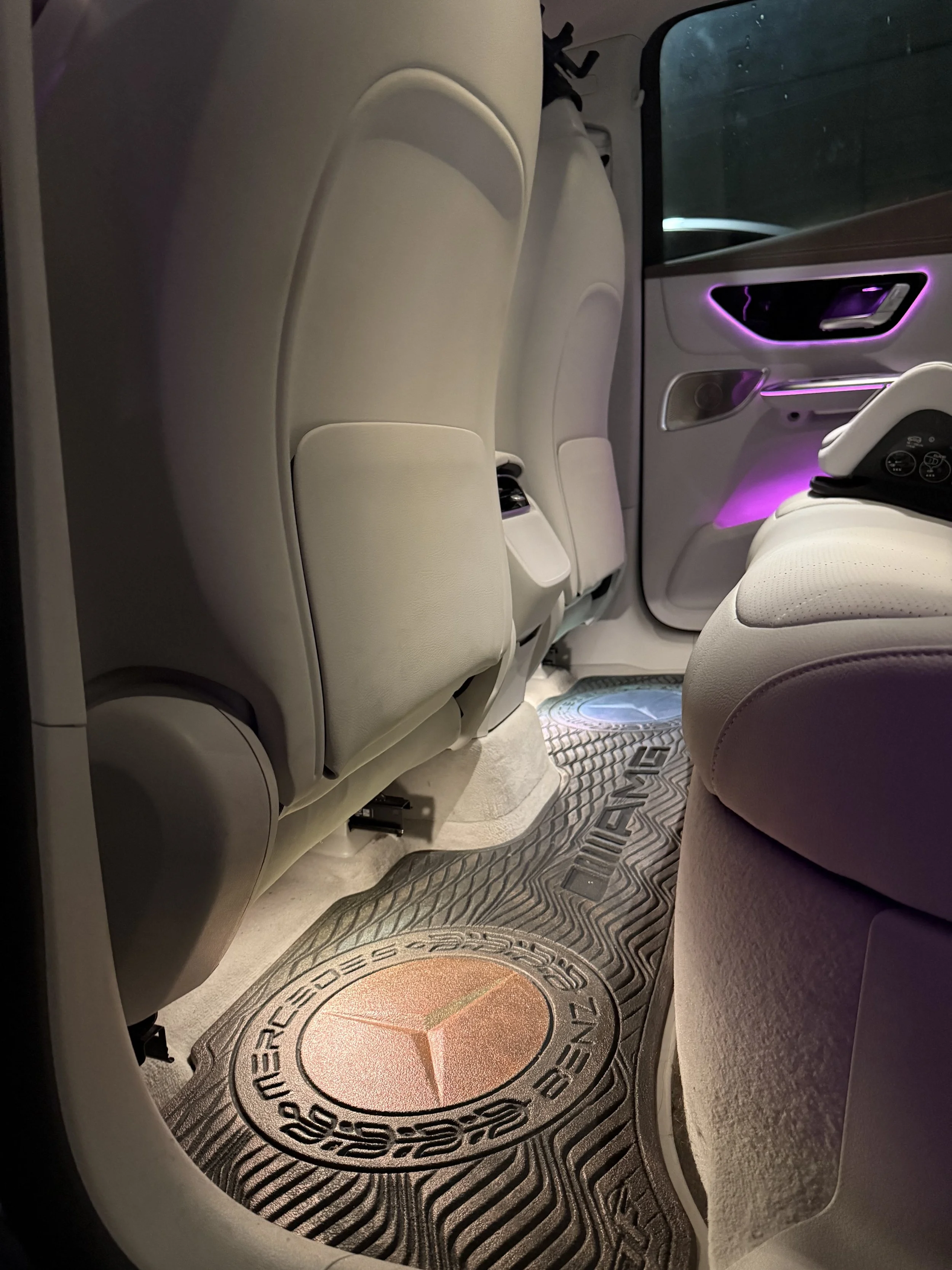Interior of a Mercedes-Benz vehicle showing beige leather seats, with features like ambient purple lighting visible on the door panel and footwell, and a custom Mercedes-Benz AutoFloor.