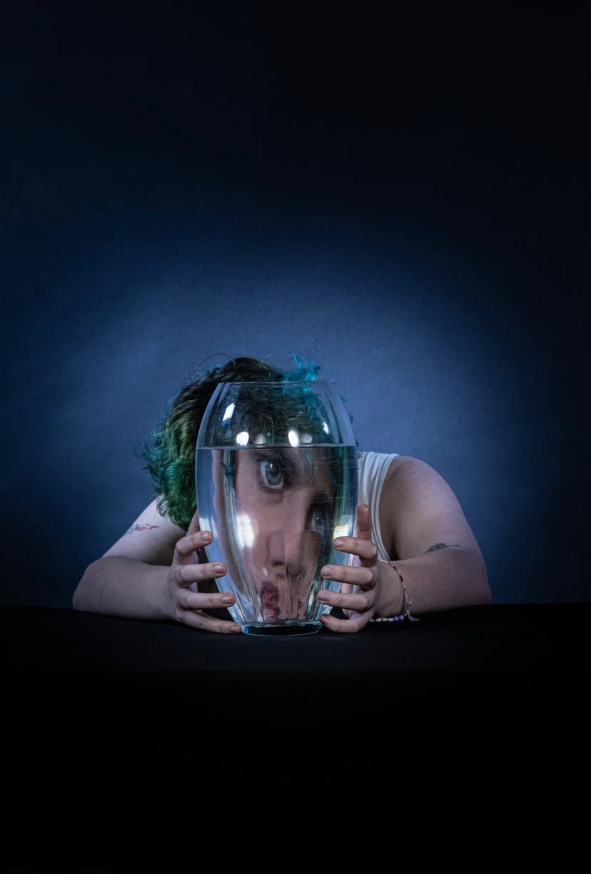 Person with green hair looking through a fishbowl filled with water with a distorted reflection, against a dark background.