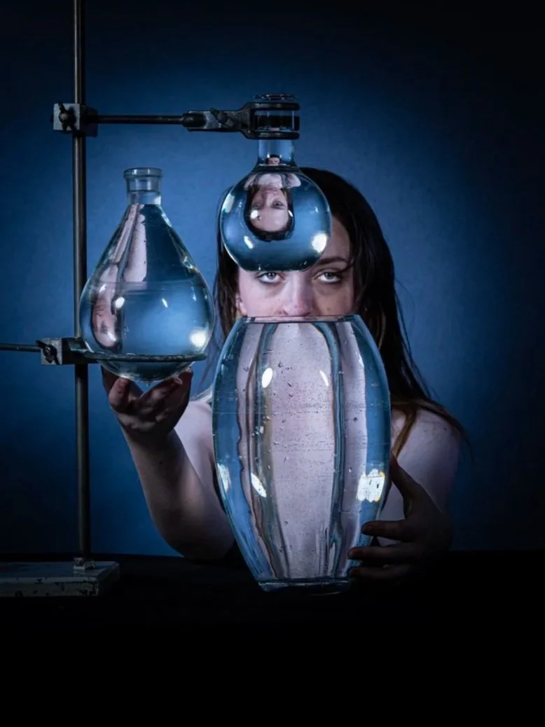 woman with dark hair looks directly at camera behind an arrangement of laboratory glassware filled with water, including a large vase and a flasks, with her face reflected in the water and upside down.