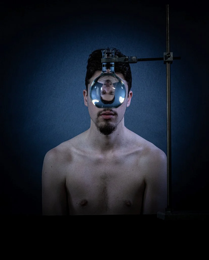 A shirtless man faces the camera with a reflective glass sphere mounted on a stand in front of his face, creating a upside-down reflection of his face within the sphere. The background is a plain dark wall.