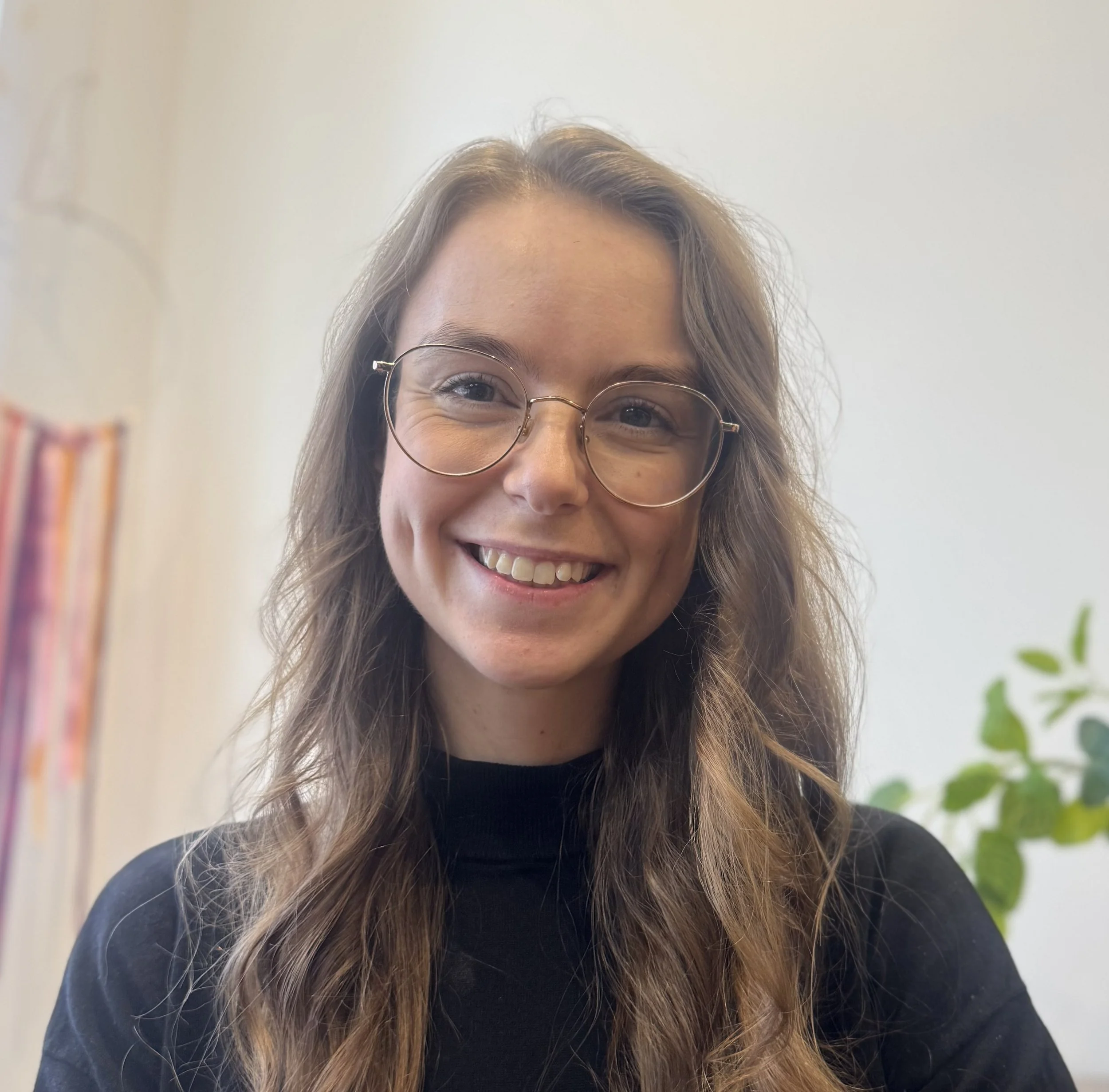 A woman with long wavy brown hair, wearing glasses and a black top, smiling in an indoor setting with a white wall and green plant in the background.