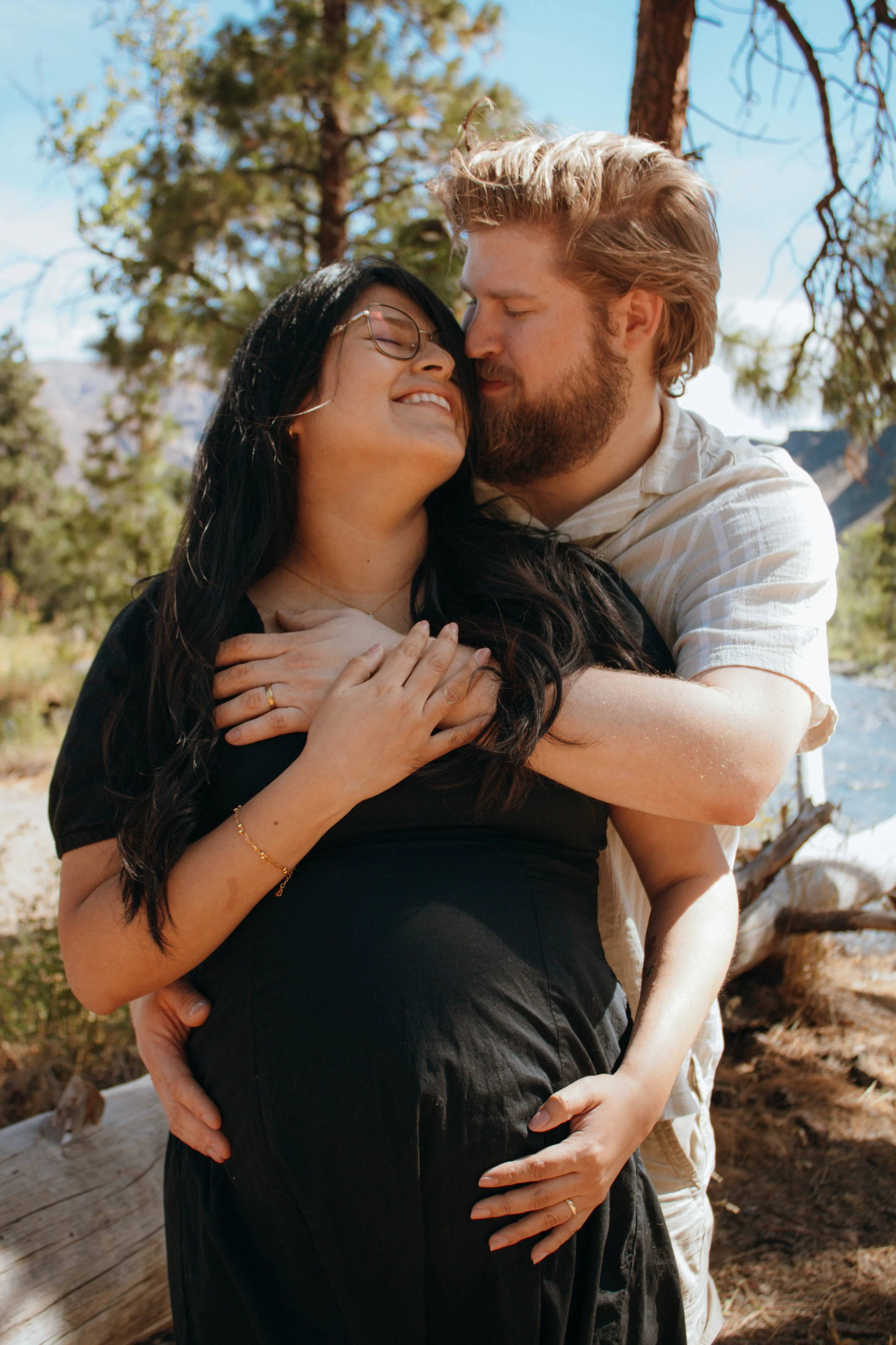A couple embracing outdoors, the woman with glasses has her hands on the man's chest while he holds her waist. They are surrounded by trees and natural scenery.