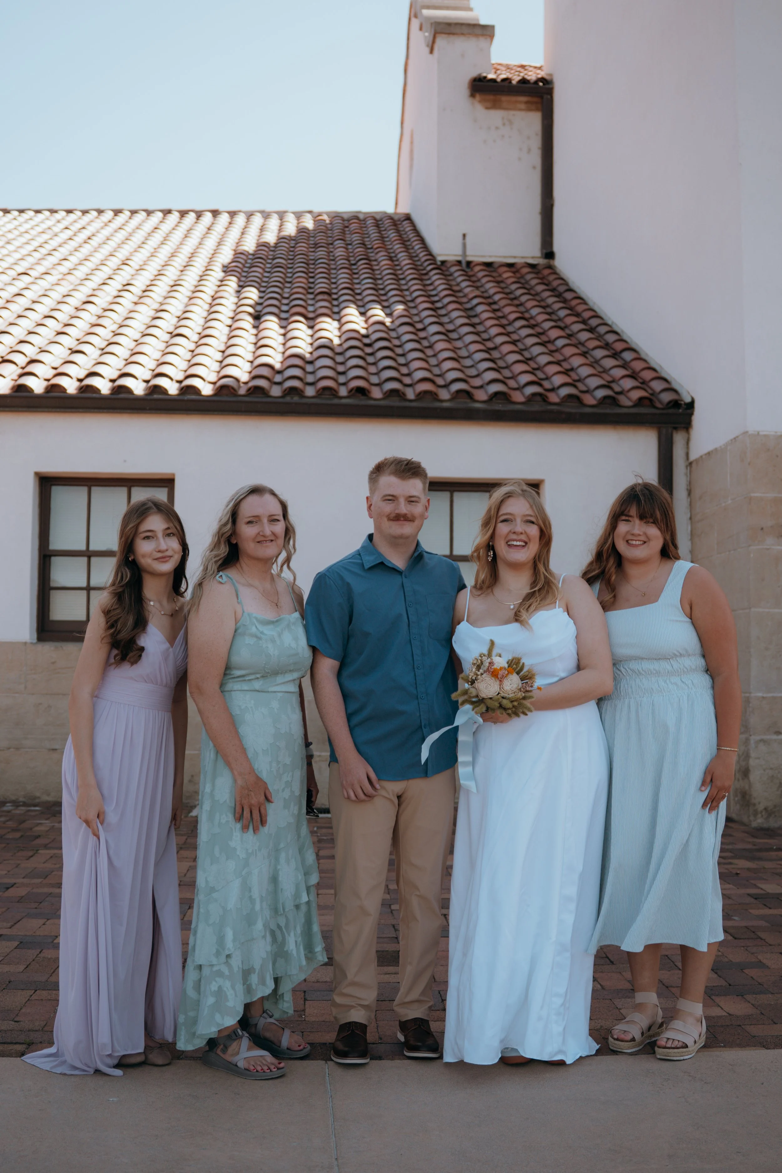 A group of five women and one man standing outside in front of a white building with a tiled roof. The women are dressed in pastel and white dresses, and one woman is holding a bouquet of flowers. They are smiling and posing for a photo under a clear blue sky.