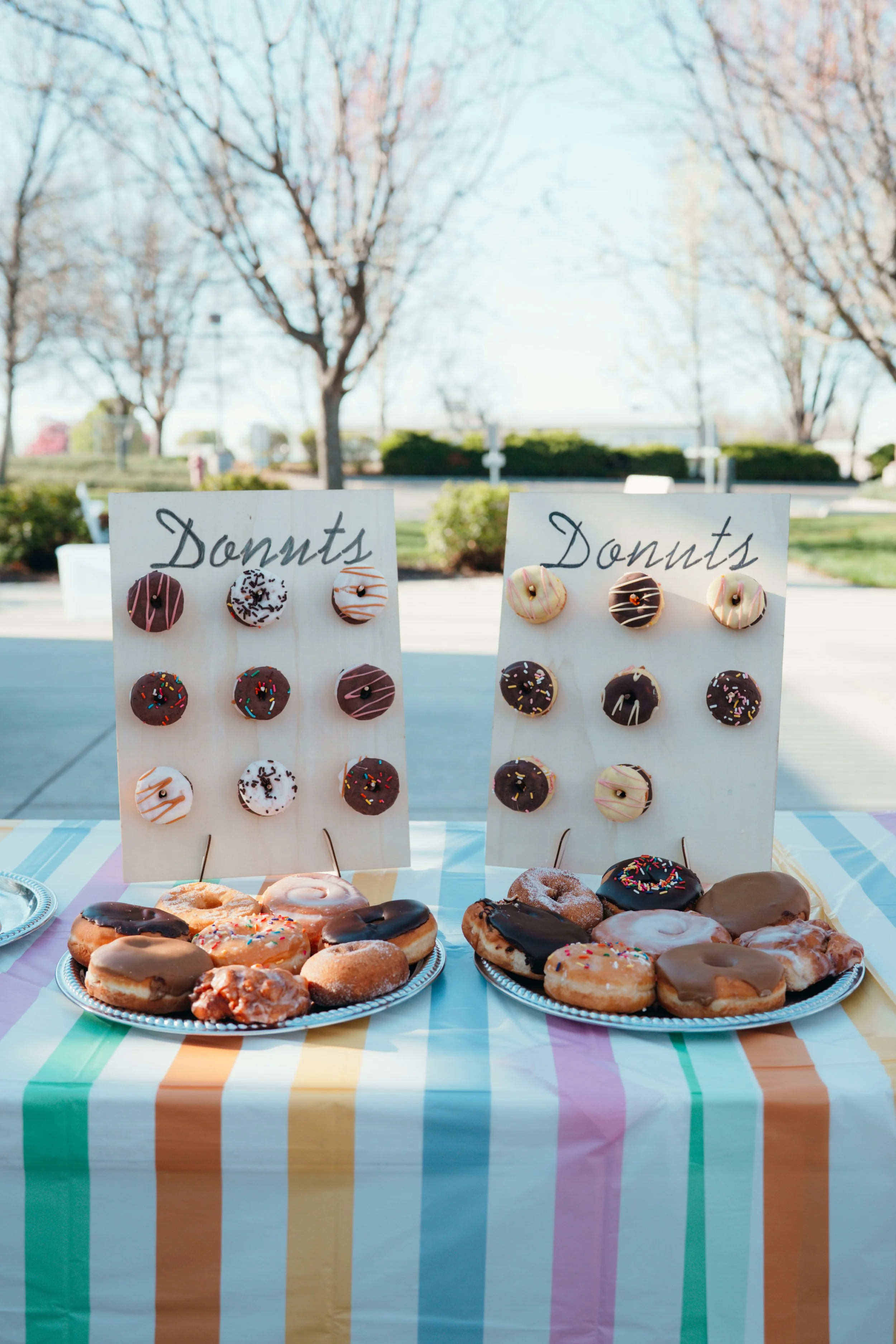 Display of assorted donuts on a table with pastel striped tablecloth outdoors. Two signs labeled 'Donuts' in cursive handwriting with decorated mini donuts attached are behind the display.