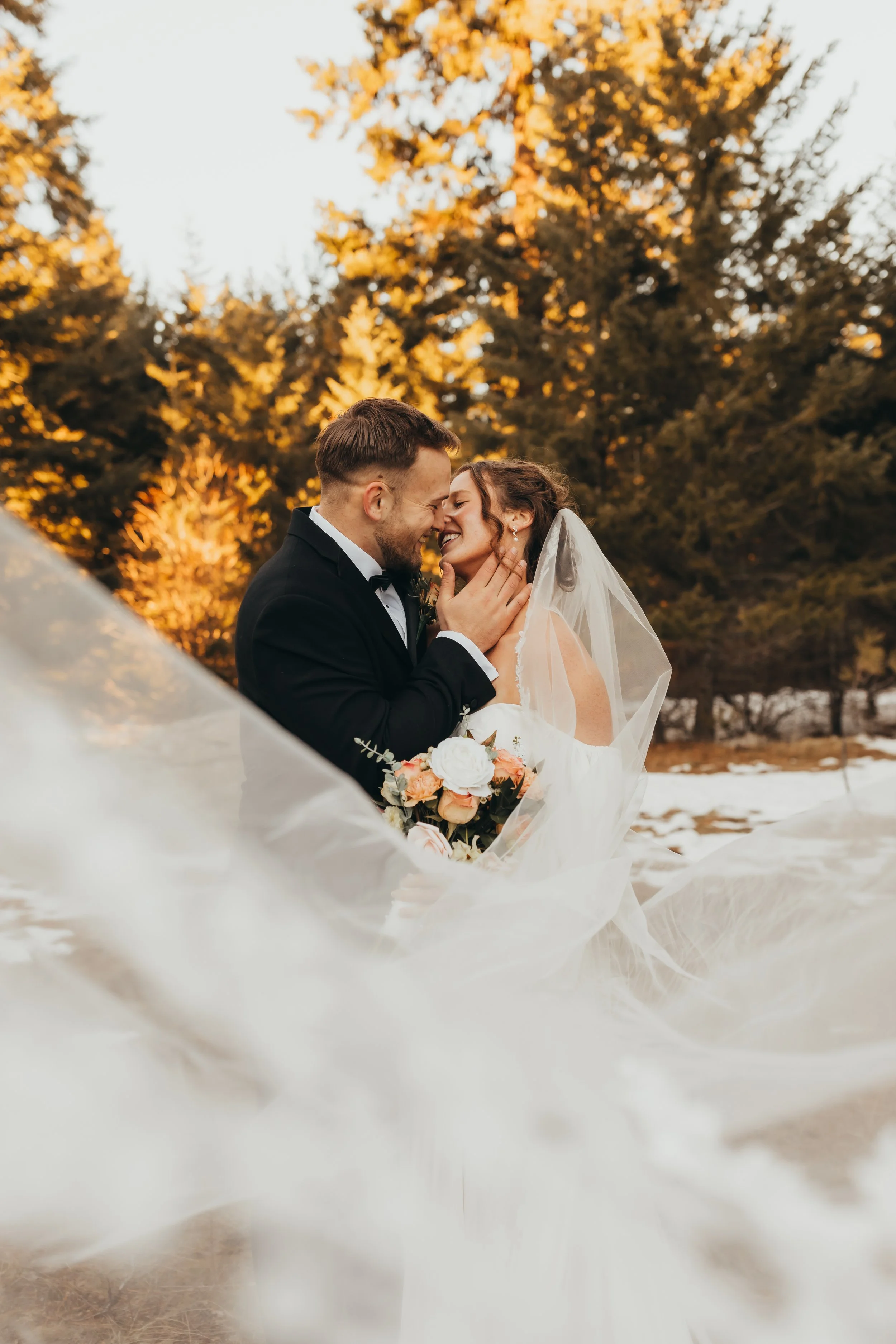 A newlywed couple sharing a kiss outdoors during sunset, with trees in the background and a white veil flowing around them.