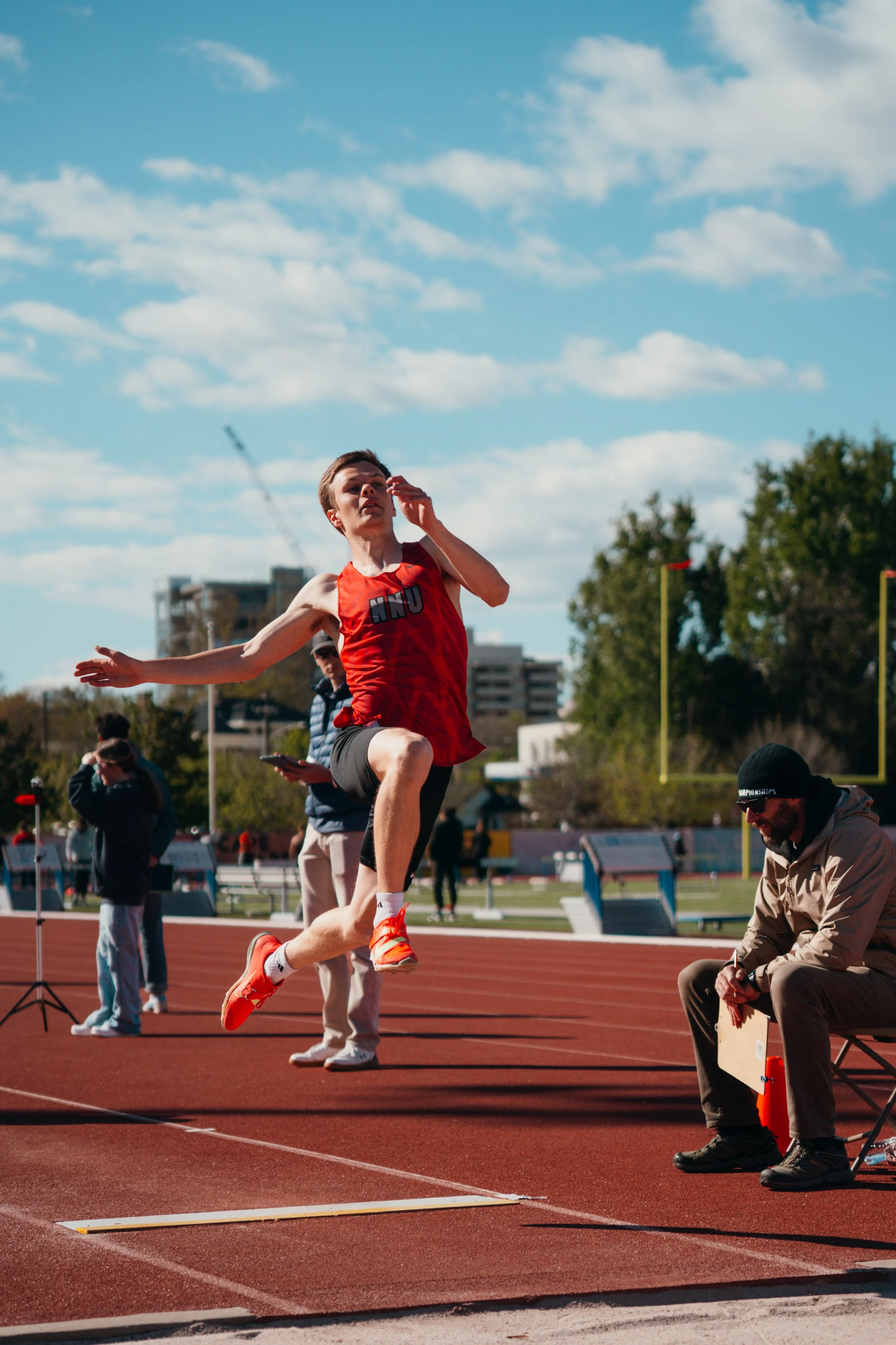 A male athlete in a red and black uniform is jumping in a track and field event, mid-air over a sand pit, with a blue sky and clouds overhead. Several people and officials are present, watching and recording the event.