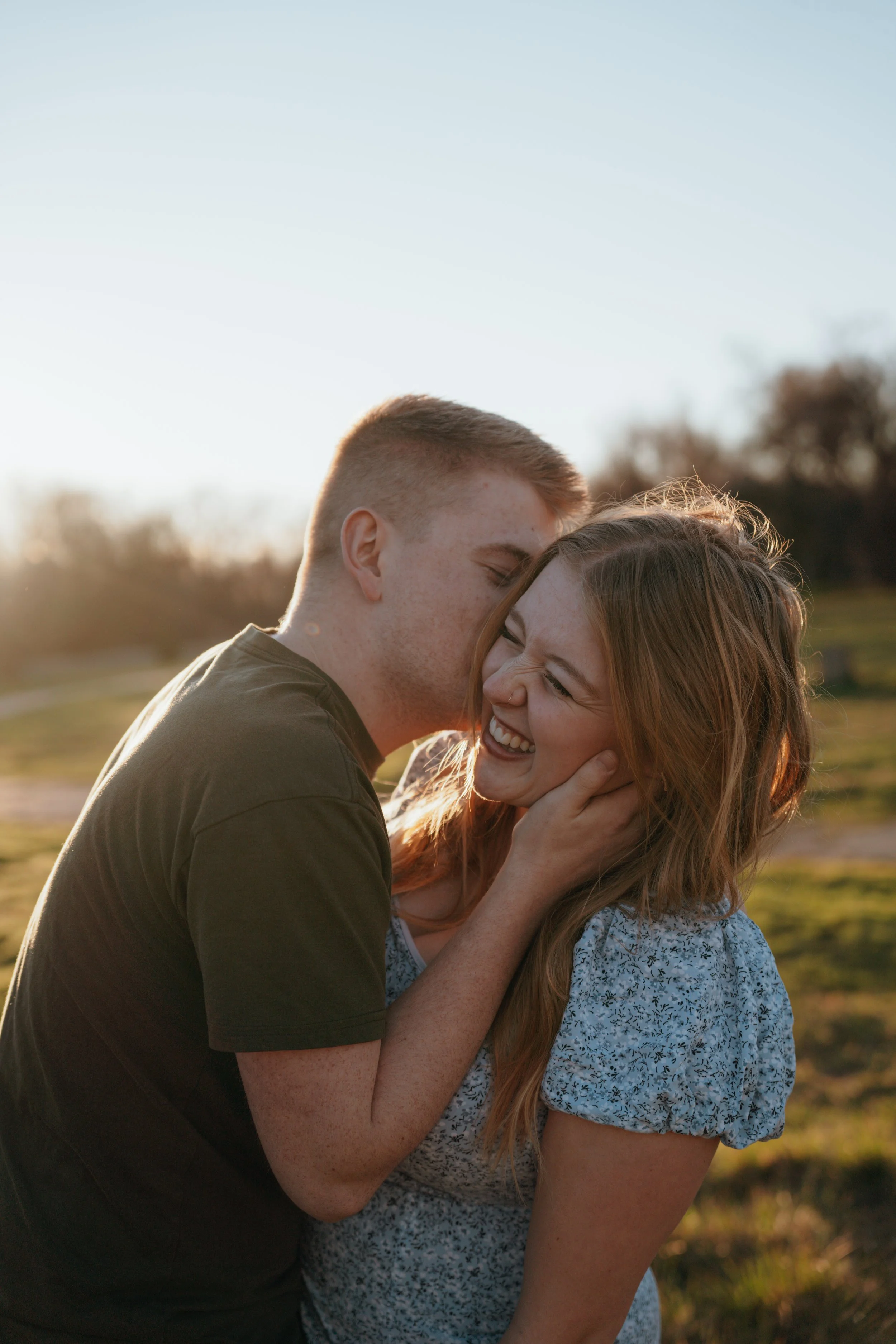 A young man and woman are outdoors, smiling and laughing as they share an intimate moment during sunset. The man gently holds the woman's face and kisses her forehead.