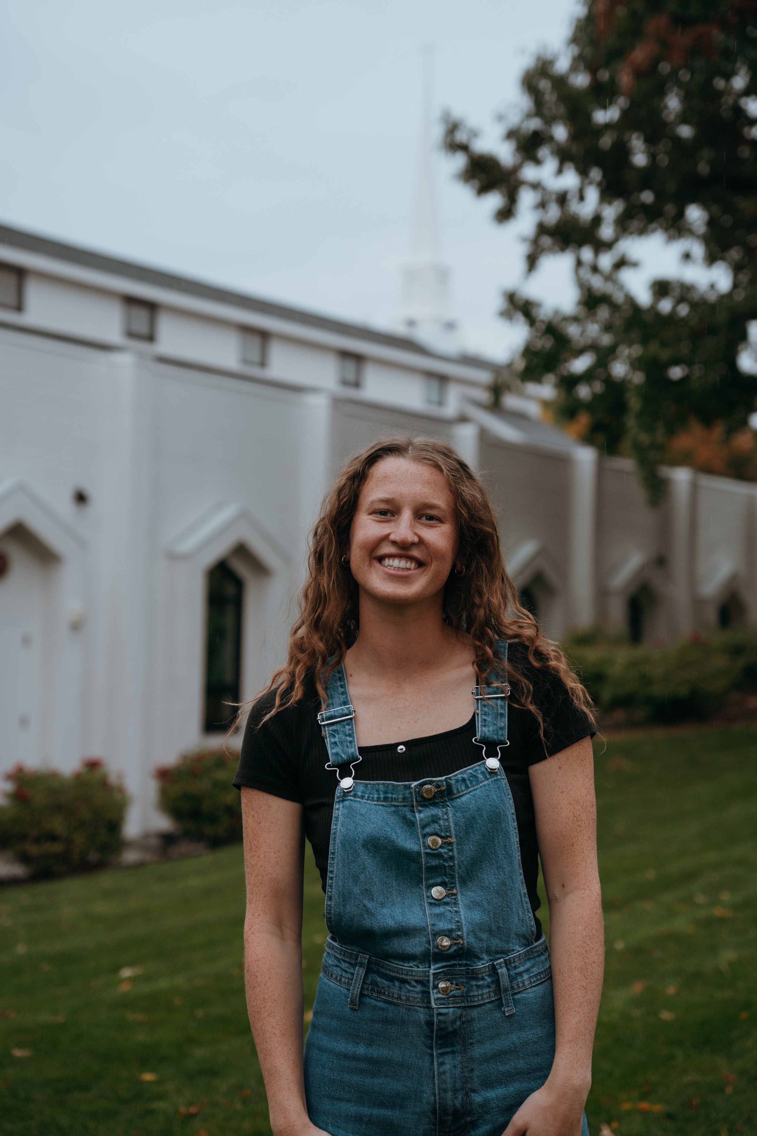 A young woman with curly red hair and freckles smiling outdoors, wearing a black shirt and denim overalls, in front of a white building and green lawn with trees.