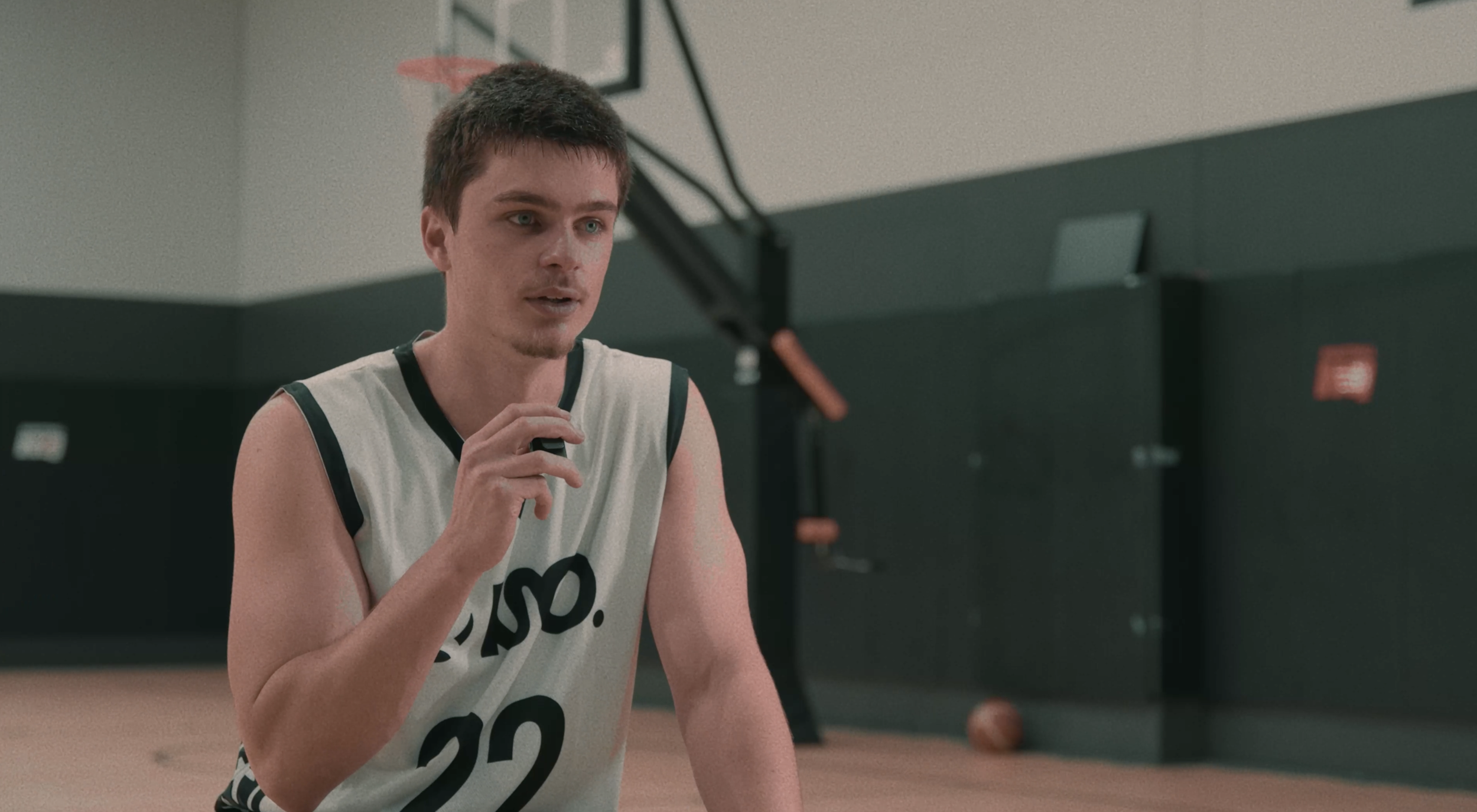 Young man in a basketball jersey speaking during a game or practice in an indoor gymnasium.