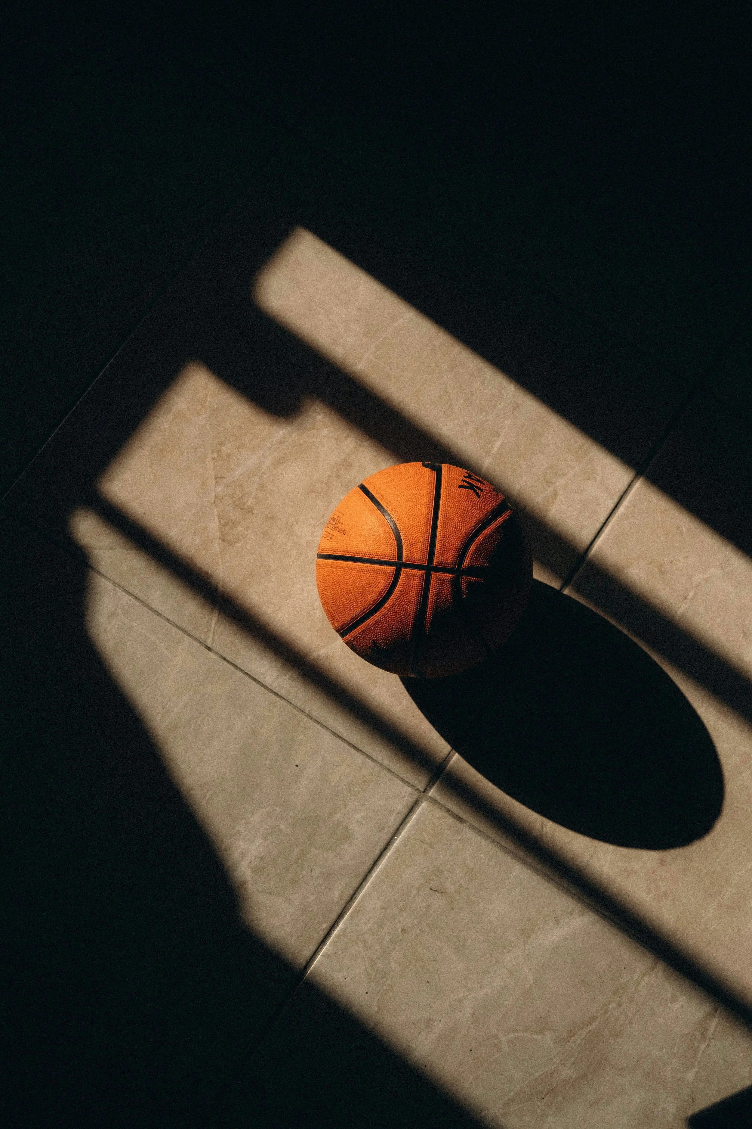 A basketball on a tiled floor, partially obscured by shadows and a dark structure.