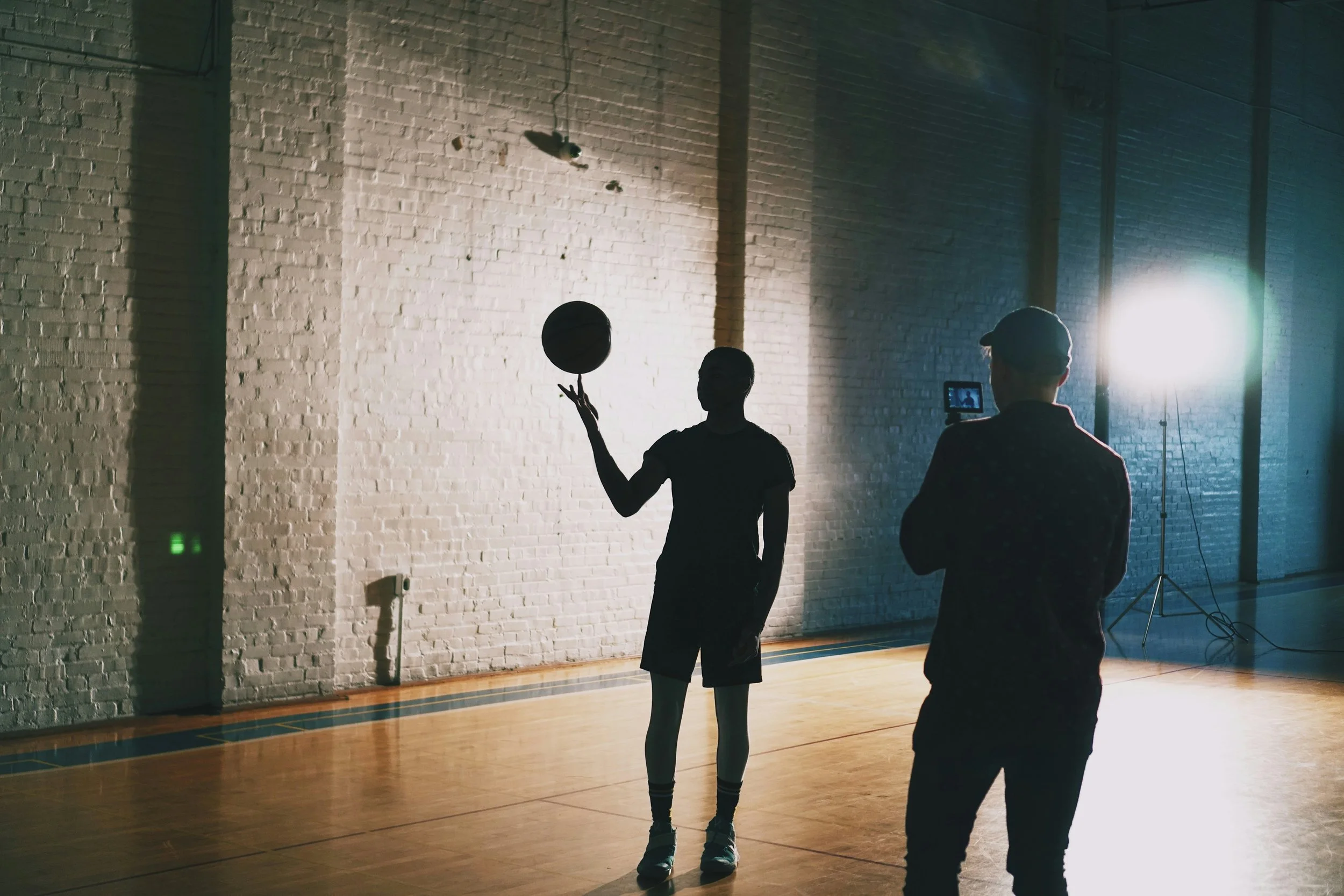 Interior of a gymnasium with a brick wall illuminated by bright lights. Two people, silhouetted, are standing on the wooden floor; one person is holding a tennis ball, and the other person is recording with a smartphone.