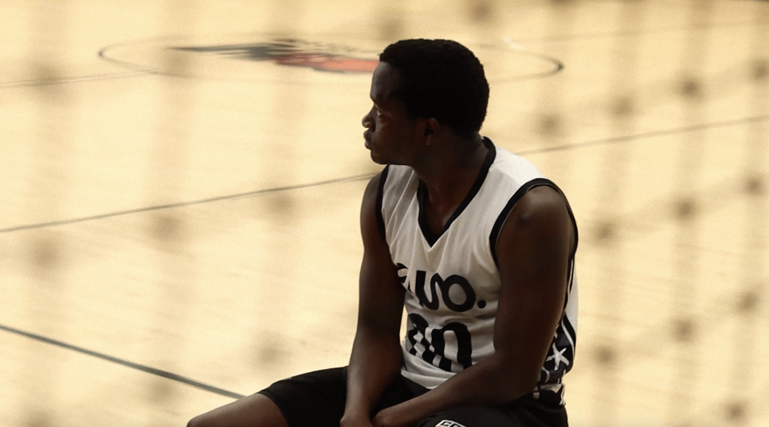 A male volleyball player sitting on the court with his eyes closed, wearing a white jersey with black accents and black shorts.