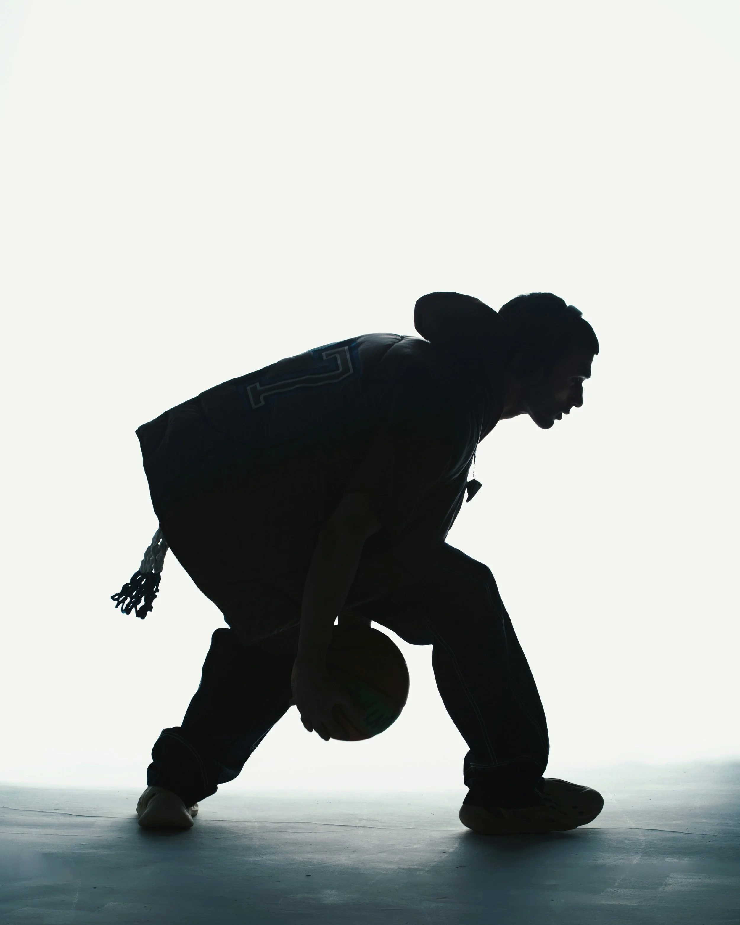 Silhouette of a person crouching with legs apart, holding a basketball on one knee, with a bright white background.