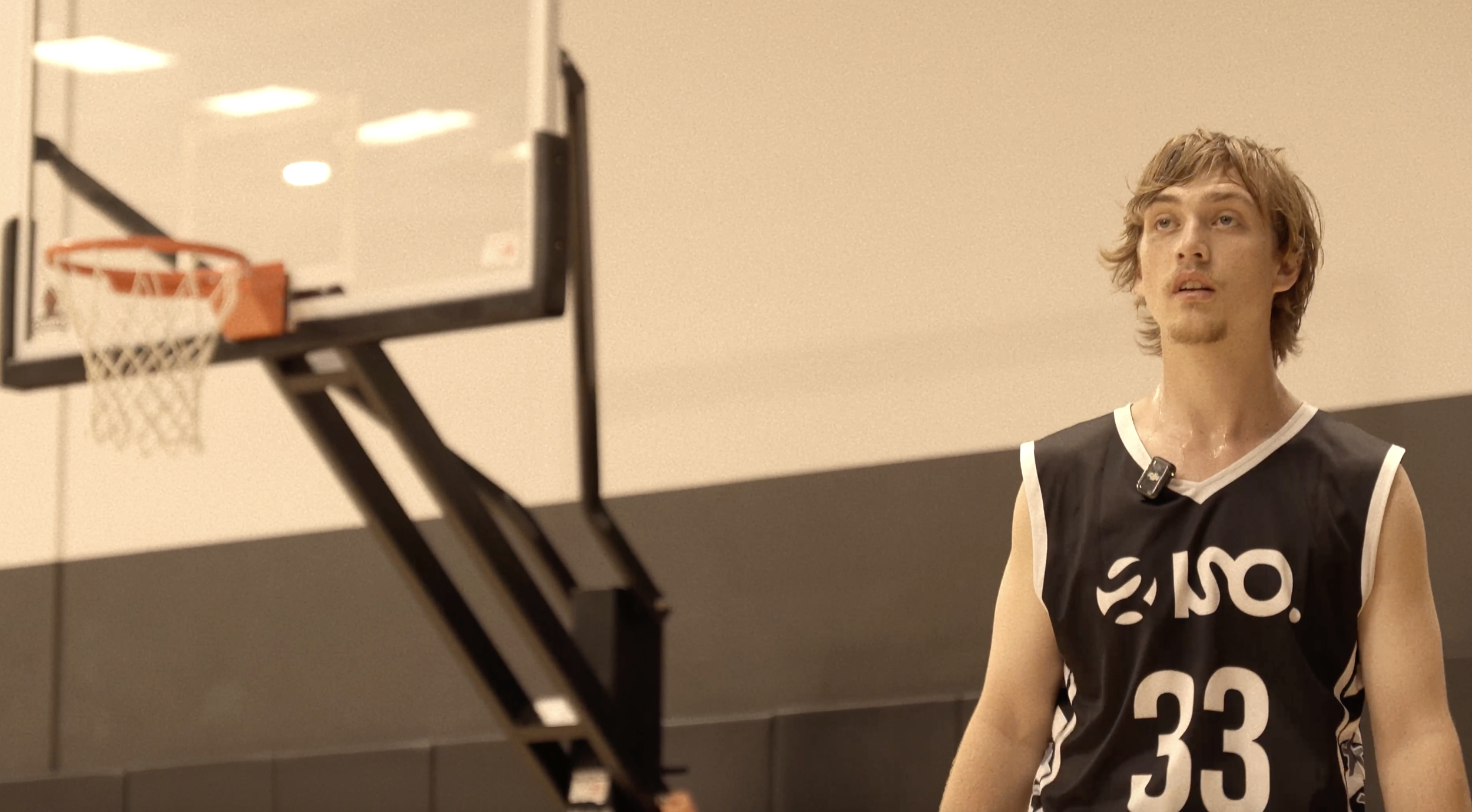 A man in a black and white basketball jersey is standing on an indoor court, with a basketball hoop and backboard visible in the foreground.