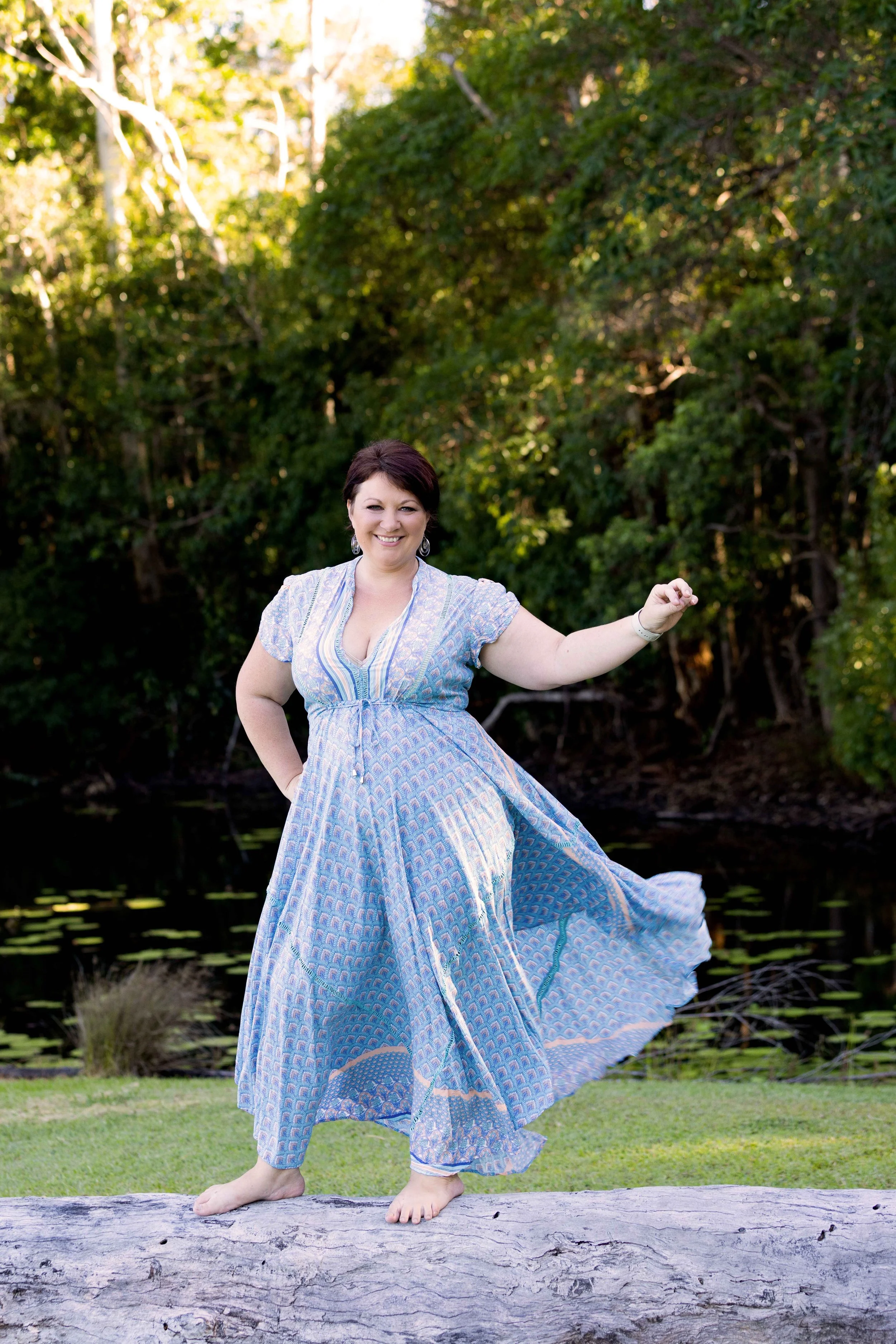 A woman with short dark hair, smiling and wearing a long patterned blue dress, standing barefoot on a log outdoors near a pond with trees in the background.
