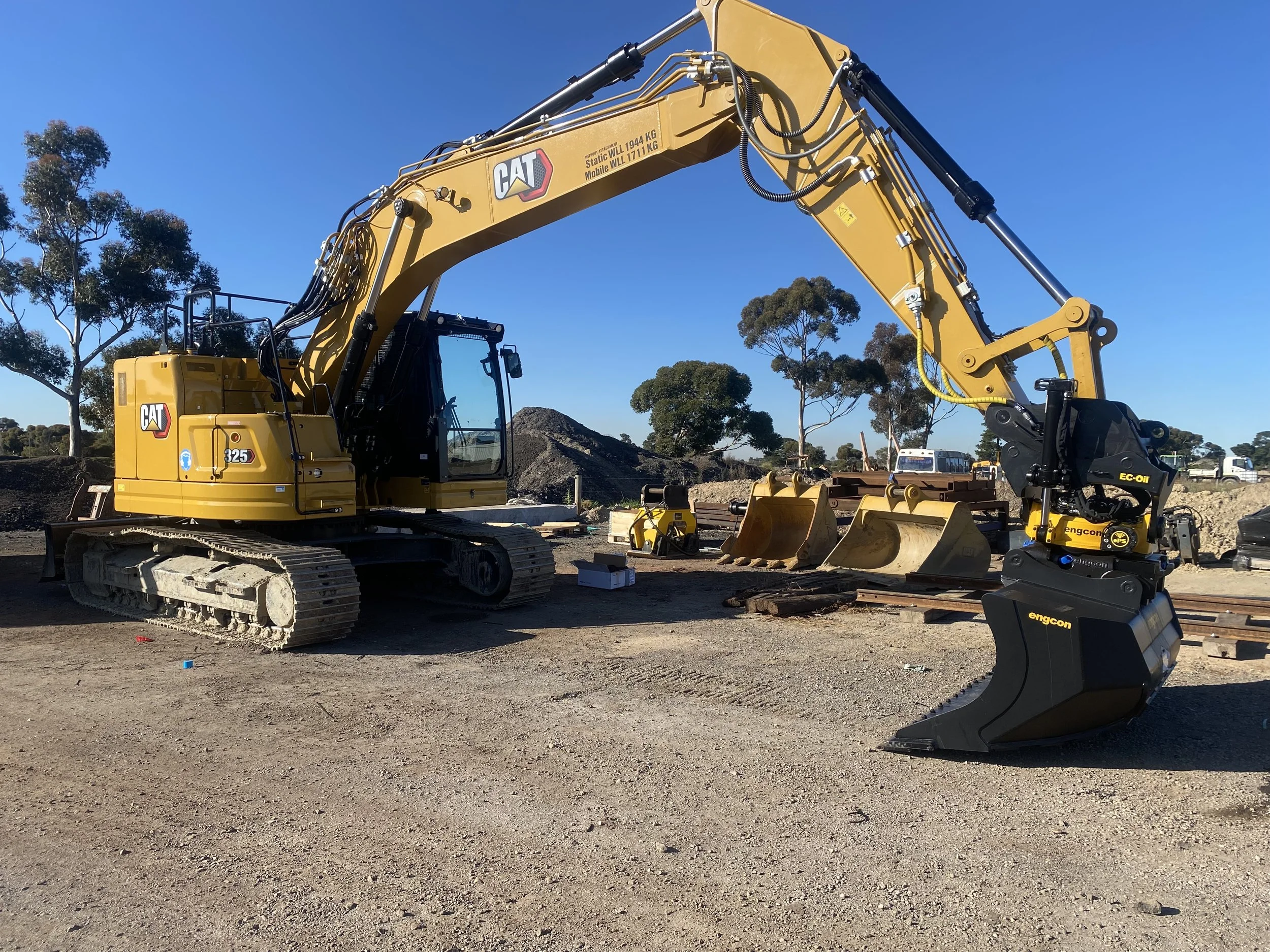 A yellow Caterpillar excavator operating on a construction site with engcon tiltrotator trees and dirt mounds in the background.