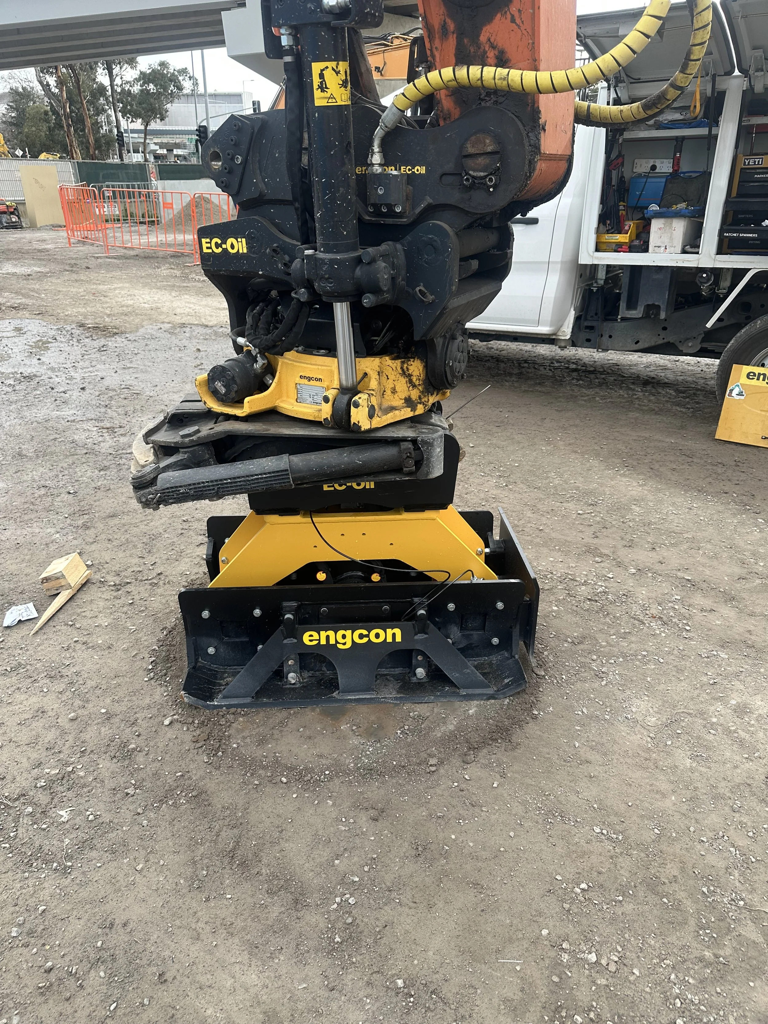A close-up photo of a engcon construction equipment attachment with the brand name 'engcon' visible on it, sitting on a dirt ground at a construction site.