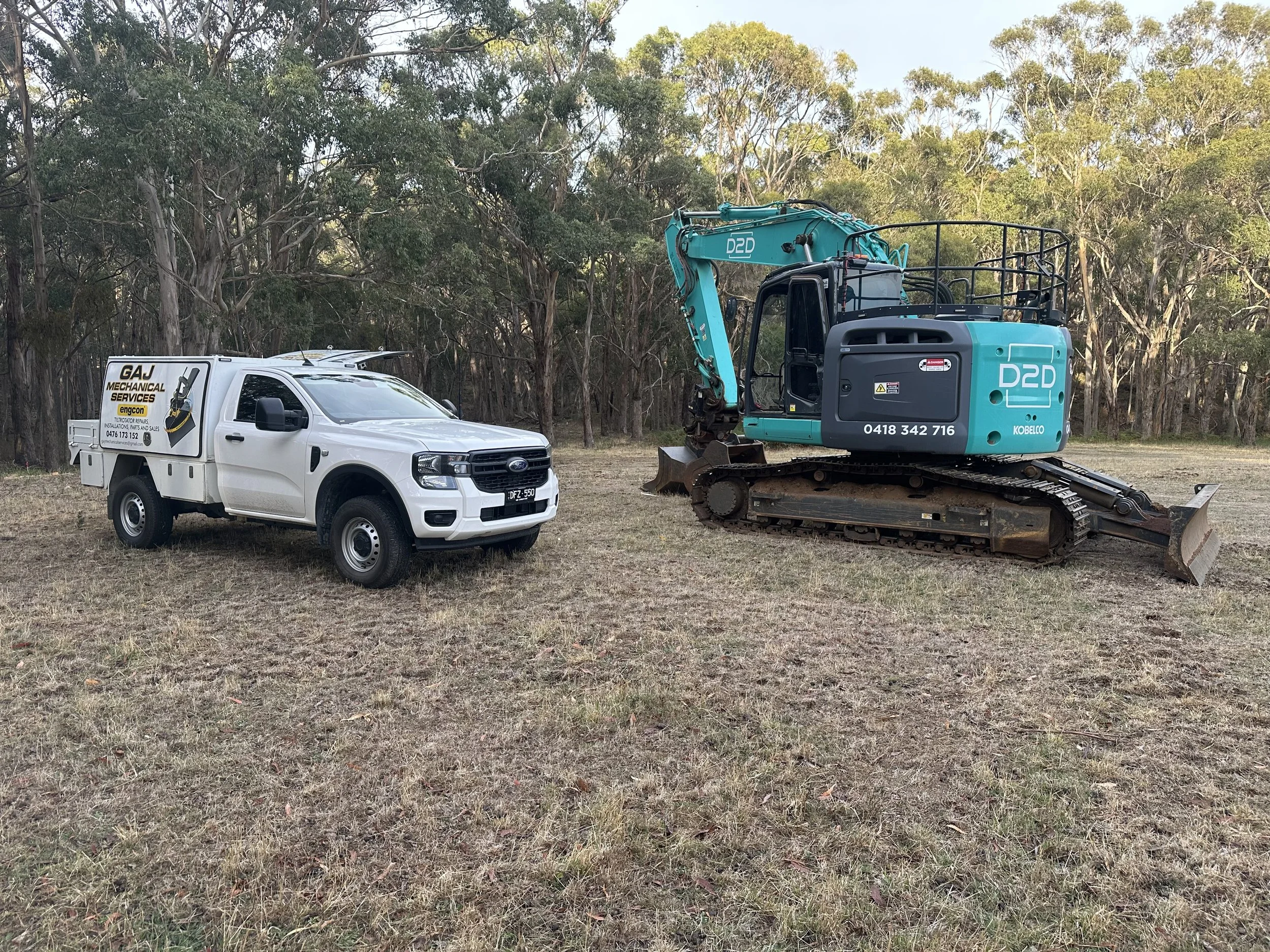 A white service truck and a blue mini excavator parked on a grassy field with trees in the background.